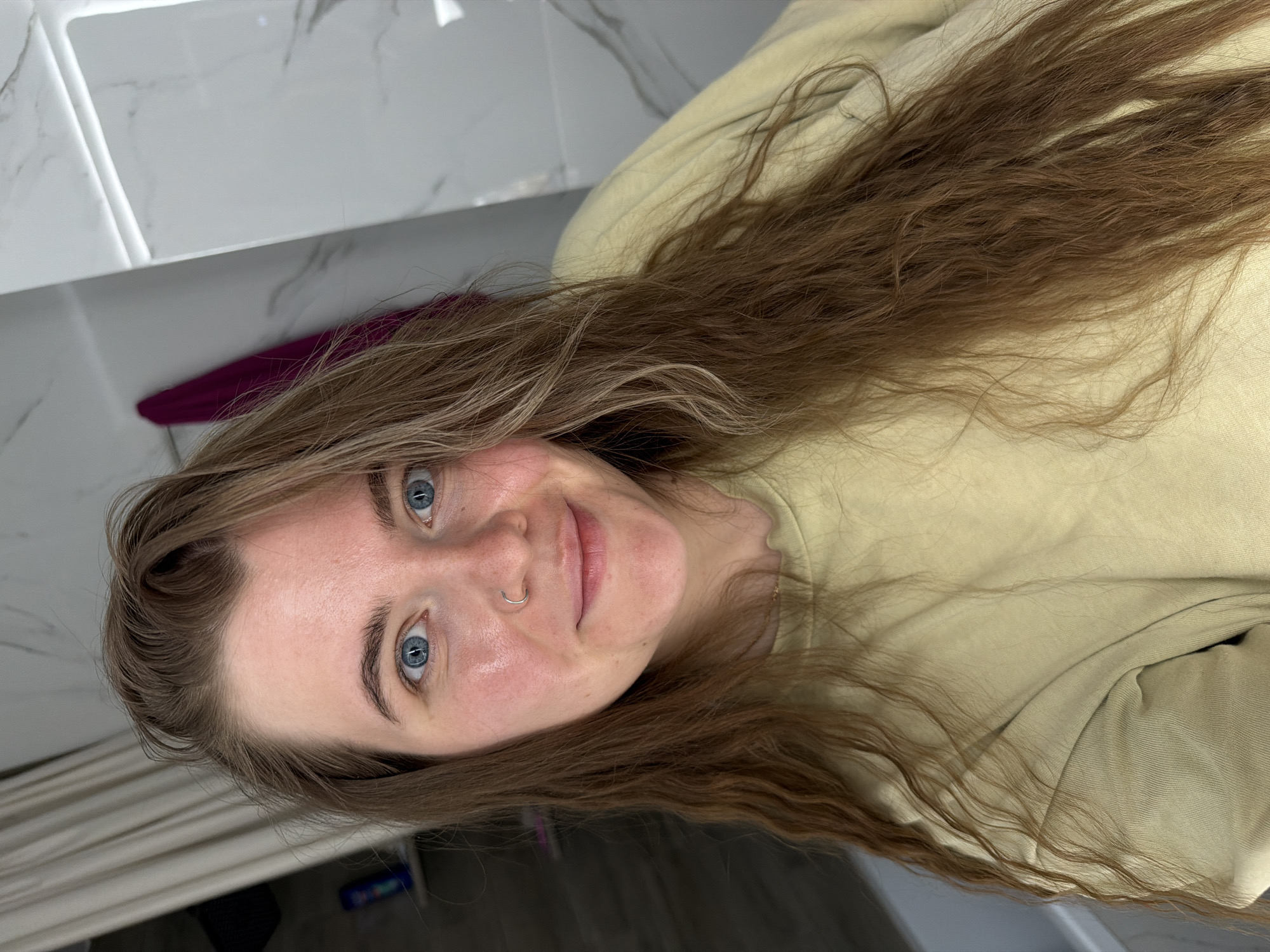 A young woman with long, wavy brown hair is centered in the frame, smiling slightly. She has fair skin, blue eyes, and a nose ring. She is wearing a light yellow top, possibly a sweatshirt or hoodie. The background consists of a white tiled wall and a dark surface below, possibly a counter. A glimpse of a purple object is visible behind her head. The photo appears to have been taken indoors. There is no information available to determine its relationship to the city of Chaikovsky, Russia.