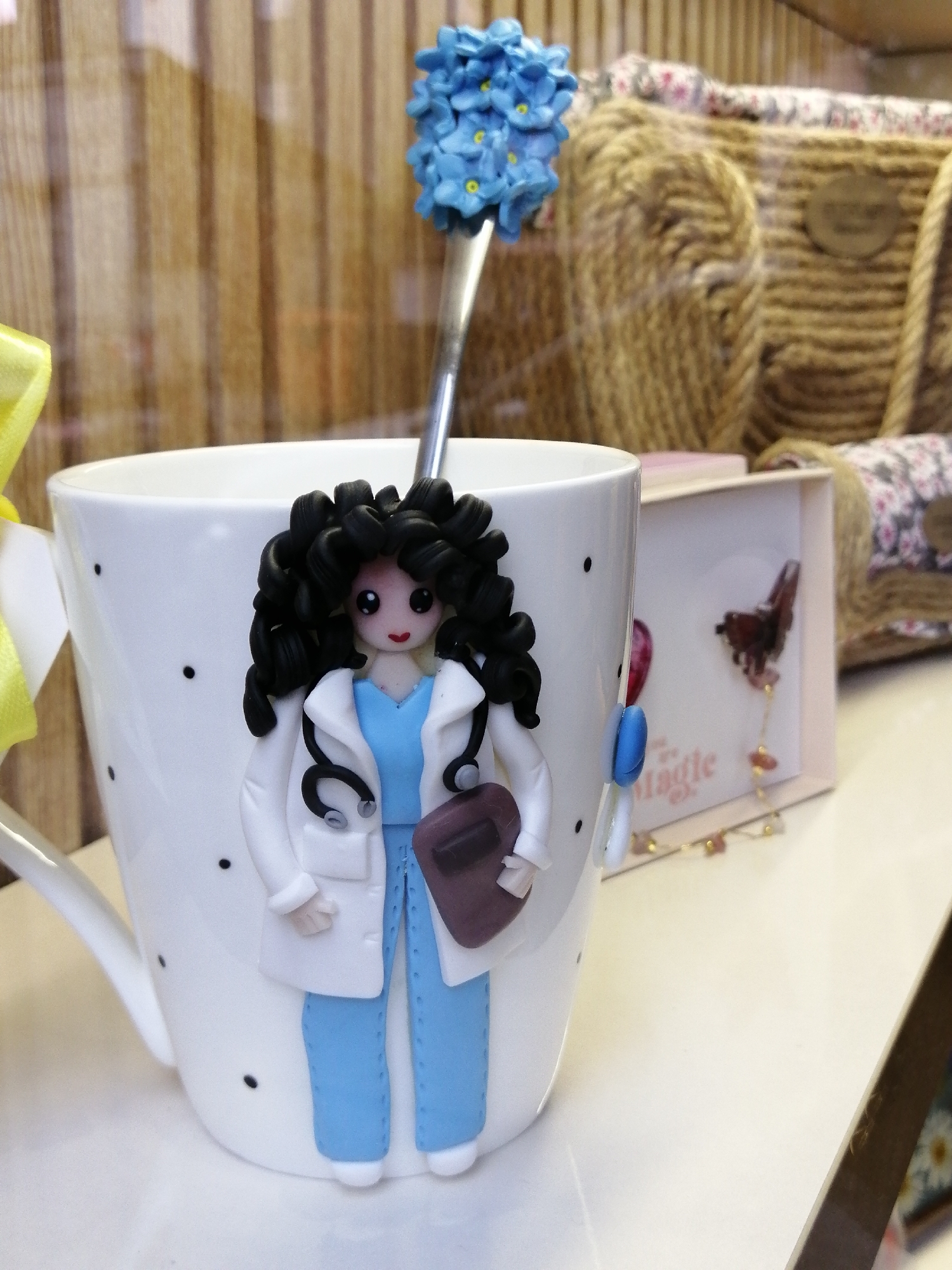 A close-up, eye-level shot captures a white ceramic mug decorated with black polka dots and a handmade polymer clay figurine of a female doctor. The doctor figure, with curly black hair, a stethoscope, a white lab coat, and blue scrubs, is affixed to the side of the mug. A silver spoon topped with a cluster of blue flowers extends from the mug, positioned behind the doctor figurine. In the background, a portion of a woven basket is visible, along with a pink box containing a decorative butterfly and beads, suggesting a retail or display setting. The overall scene is lit, with a wooden-paneled background. The photograph does not provide any visual information to determine its specific location within the city of Batumi, Georgia.