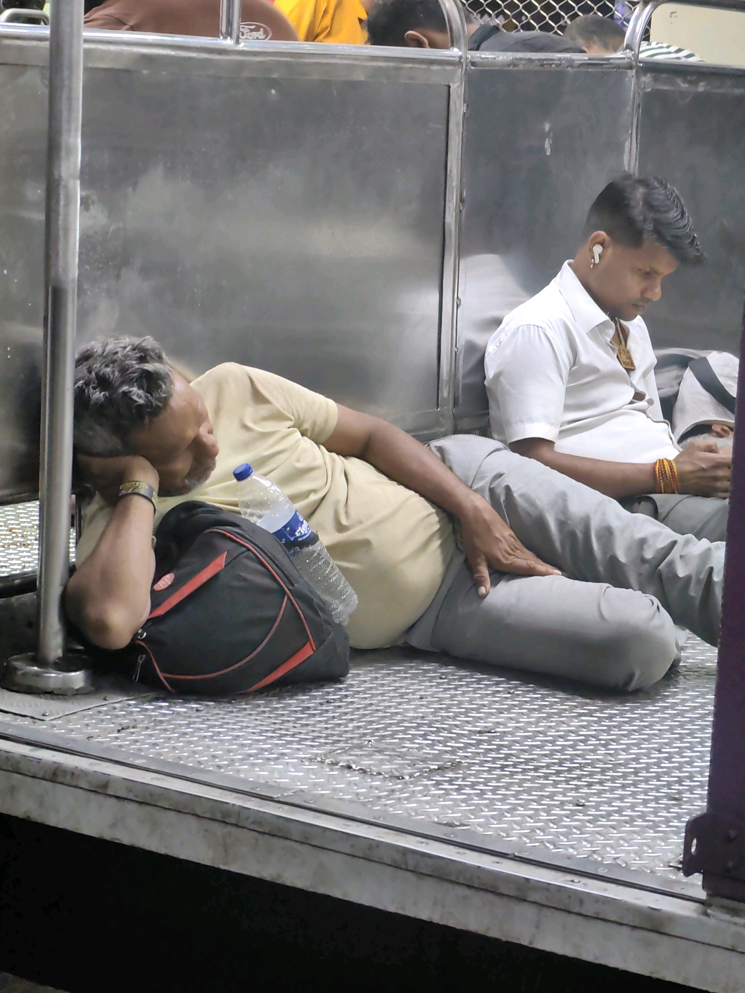 In the city of Mira Bhayandar, India, two men are visible inside a train carriage. On the left, an older man with graying hair is asleep, leaning his head on his arm and resting against a black and red backpack. He is wearing a light-colored t-shirt and appears to have a bracelet on his left wrist. Next to him, to the right, a younger man in a white shirt with ear buds in his ears is looking down. He has a necklace with a pendant around his neck and colorful bracelets on his right wrist. The carriage interior features metal walls and a textured metal floor. A plastic bottle of water is placed near the sleeping man.