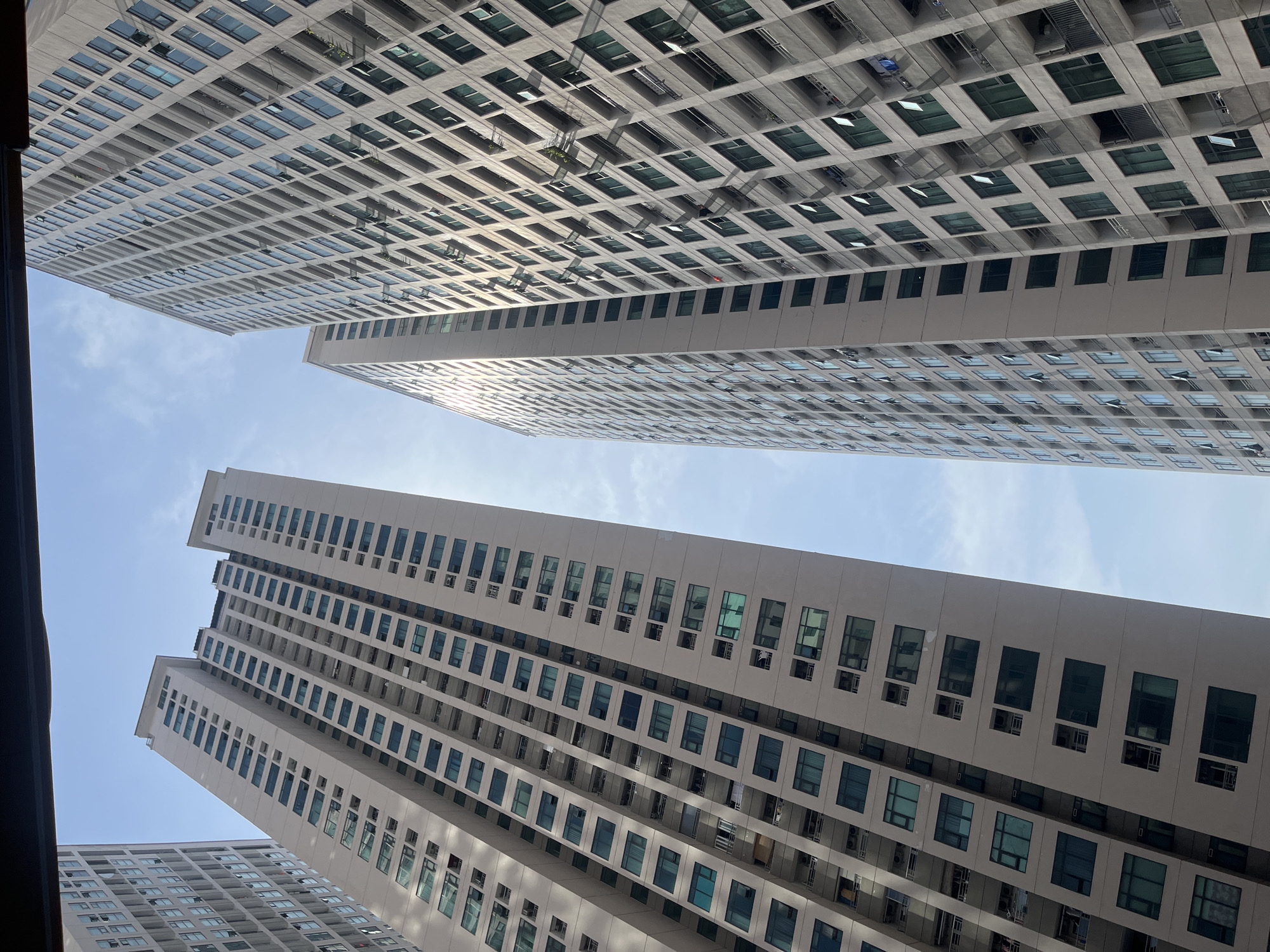 The image shows a view looking up at several very tall, modern apartment buildings against a bright blue sky with sparse clouds. The buildings are clad in a light-colored material, possibly concrete or stucco, and are characterized by numerous windows arranged in a grid pattern. Some windows are dark, reflecting the sky, while others appear to be open, revealing balconies and glimpses of the interior. The architecture suggests a dense urban environment. The perspective is from a low angle, emphasizing the immense height of the structures. The lighting indicates it is daytime, with sunlight casting shadows and highlighting the textures of the buildings. There are no visible people in the image. The overall impression is one of a contemporary cityscape with residential high-rises. The image is likely taken from within a city, given the presence of such large apartment complexes.