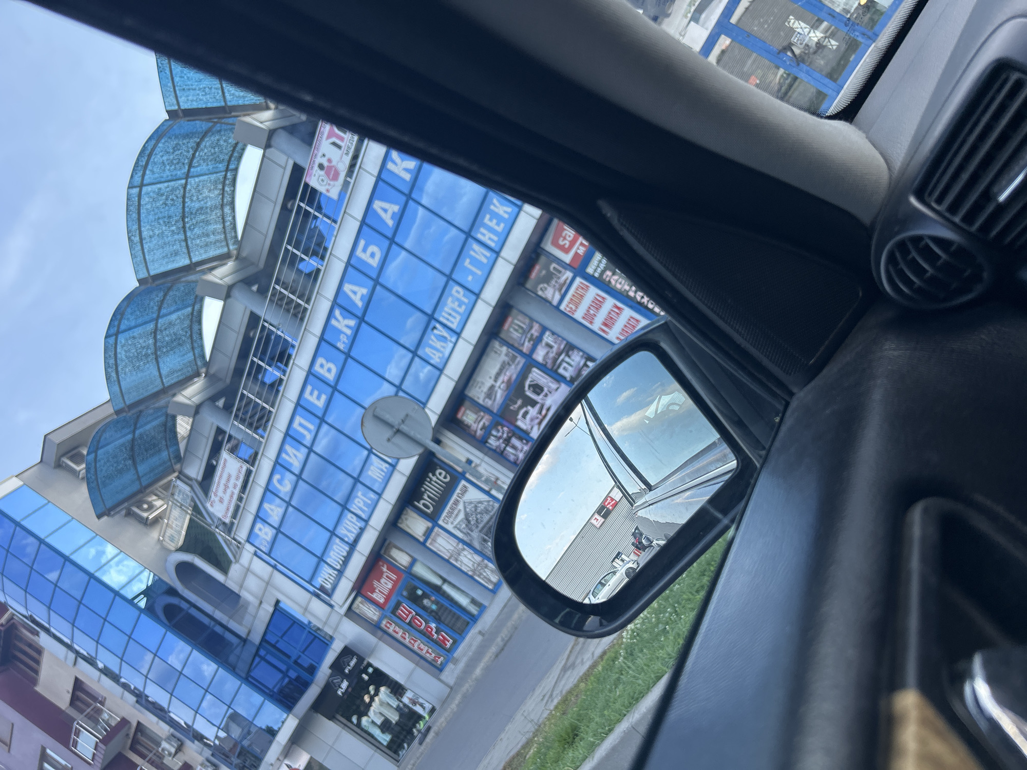This is a low-angle, eye-level, full shot captured from inside a vehicle, looking out through the driver's side window and side mirror. The foreground is dominated by the interior of the car, specifically the dashboard with an air vent and a portion of the door panel. The side mirror reflects a street scene with cars parked in front of a modern building. The building is tall and features a blue glass facade with several visible signs in a Cyrillic alphabet. One prominent sign reads "ВАСИЛЕВ-КАБА" in large blue letters. Below this, another sign says "АКУШЕР - ГИНЕК". Other smaller signs for businesses like "brilliant" and "brillife" are also visible. The building has a blue, curved canopy structure on its upper levels. The environment appears to be an urban setting during daylight.