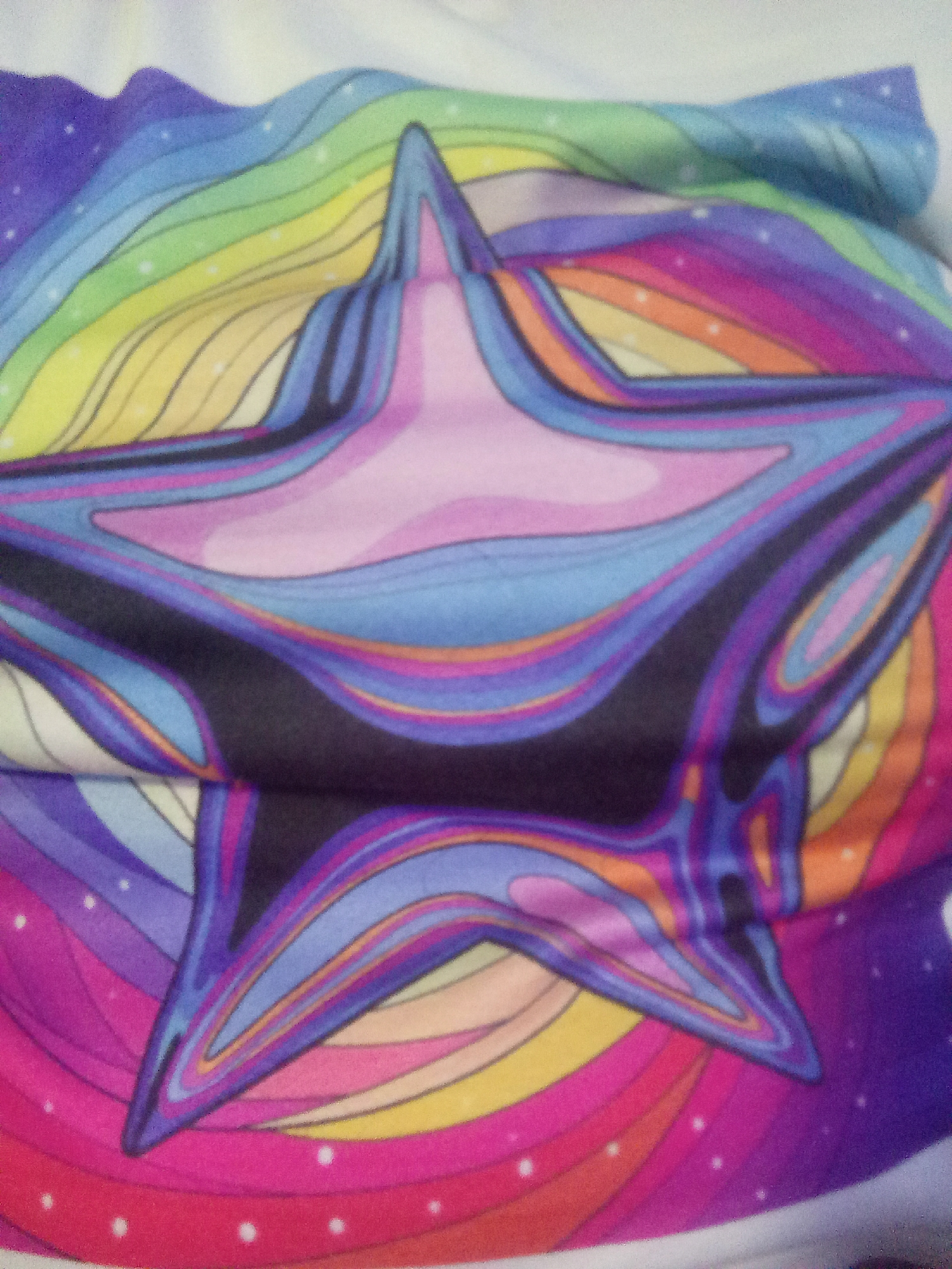 The image shows a close-up, slightly blurry view of a colorful star-shaped design printed on fabric. The design features a central pink and light blue star with thick, wavy outlines in black, dark blue, purple, and orange. Surrounding the star is a rainbow-colored pattern with curved lines that radiate outwards, transitioning through purple, blue, green, yellow, orange, and red. Small white dots, resembling stars or sparkles, are scattered across the purple and pink sections of the background. The fabric appears to be folded or creased in places, causing distortions in the pattern. There is no discernible background or environmental context that would indicate a specific location in La Tagua, Venezuela. The image is a still shot and does not capture any actions or interactions.