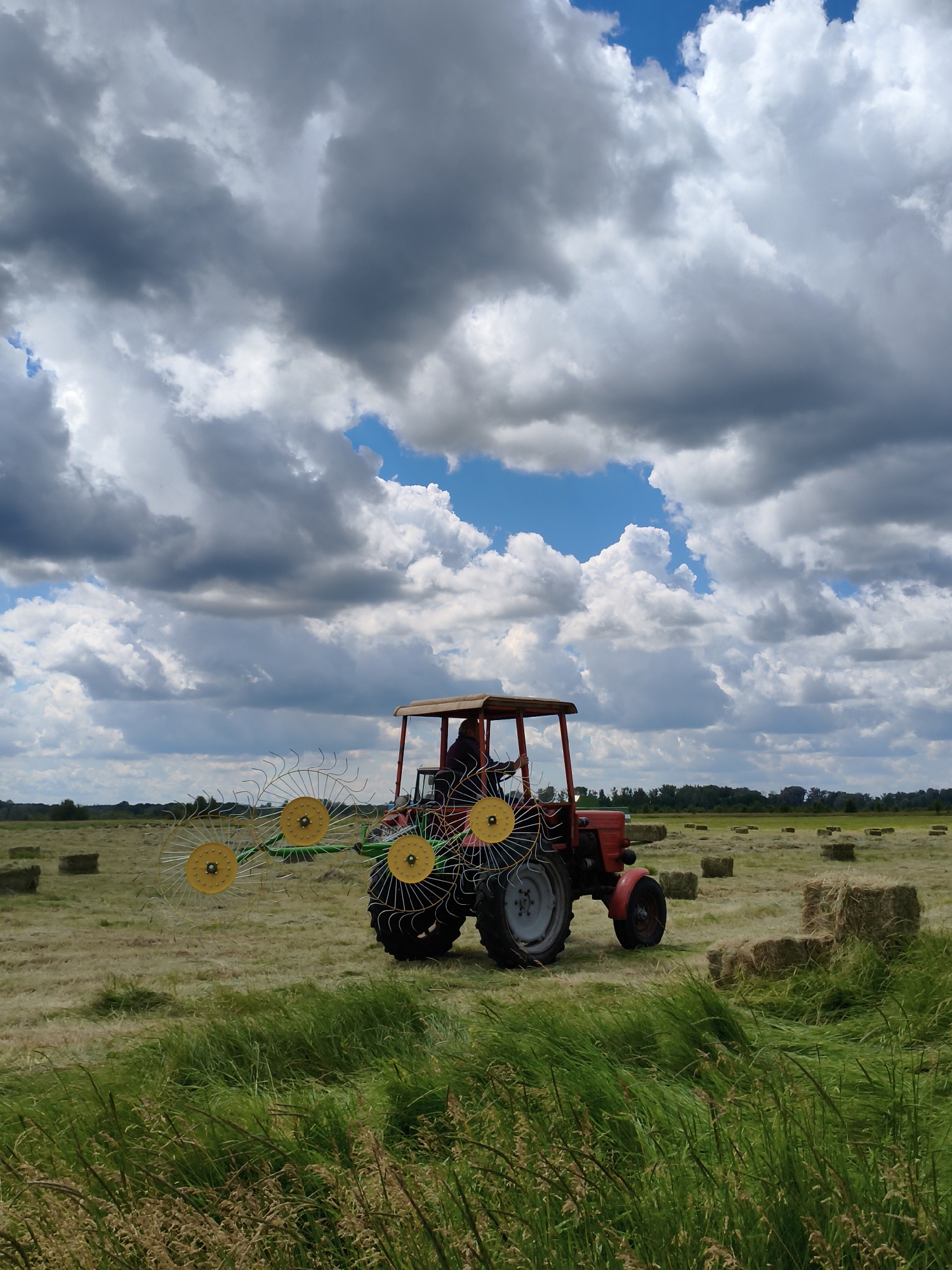 In Ustyvytsia, Ukraine, a red tractor with a hay rake attachment is operating in a field of cut hay. The tractor is positioned in the center of the frame, and its rake is extended to the left, featuring several large, yellow-centered circular components with wire tines. A person is seated in the driver's seat of the tractor. The field is dotted with numerous rectangular bales of hay, some stacked and others individually placed. The foreground is dominated by tall green grass. The sky above is dynamic, with a mix of dark, heavy clouds and patches of bright blue sky. The background shows a treeline in the distance.