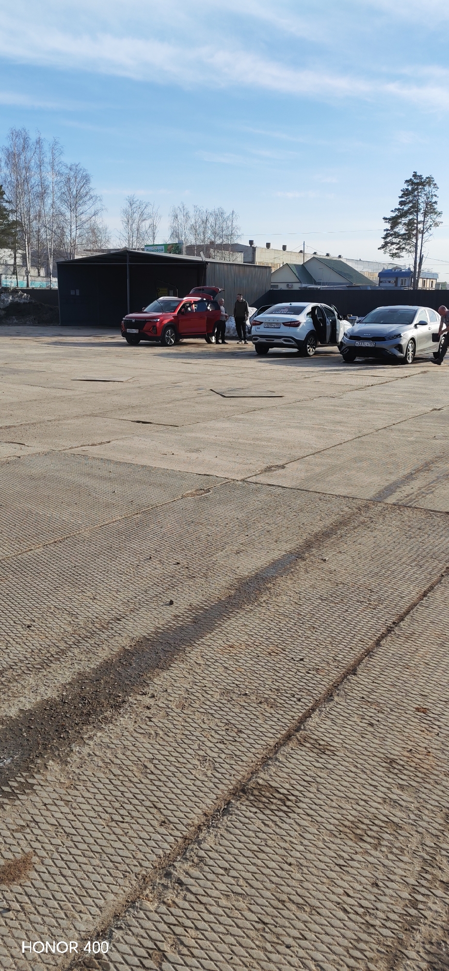 The image shows a parking area in Chaikovsky, Russia on a sunny day. The area is paved with textured concrete slabs. In the background, there are several buildings, some bare trees, and a single evergreen. In the parking area, there are three cars: a red Changan, a white Lada, and a gray Kia. The trunks of the red and white cars are open. Several people stand around the vehicles. The photo has a watermark "HONOR 400" in the lower left corner.