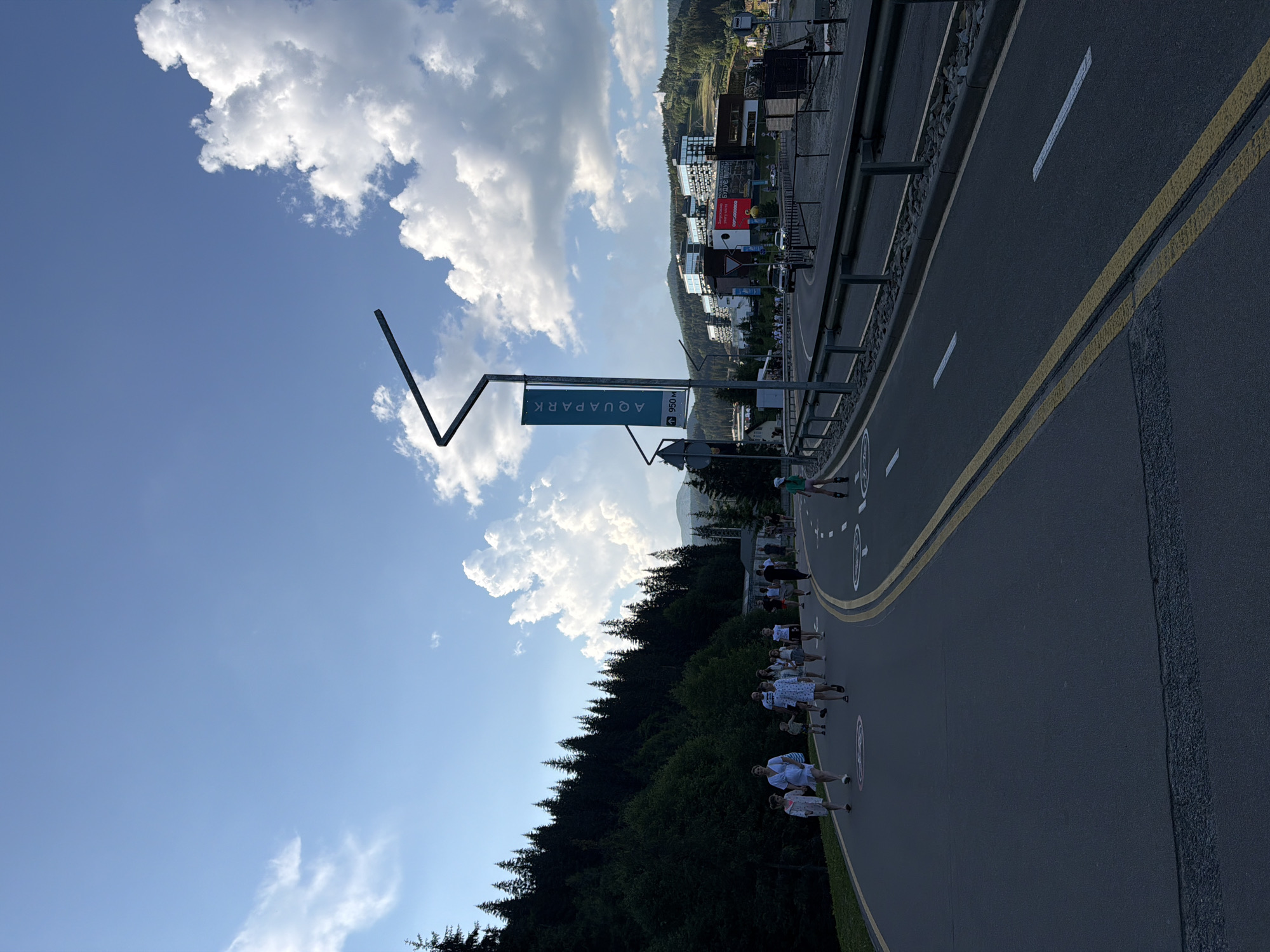 A group of people are walking along a paved path next to a road lined with yellow markings. In the background, there are buildings nestled on a hillside, surrounded by trees. The sky is blue with scattered white clouds. A signpost in the foreground reads "AQUAPARK" with an arrow pointing to the right and "056 km" below it. The scene appears to be outdoors in a mountainous or hilly area, possibly during daytime. The people are dressed casually, and their activity suggests a leisurely walk or a group outing.
