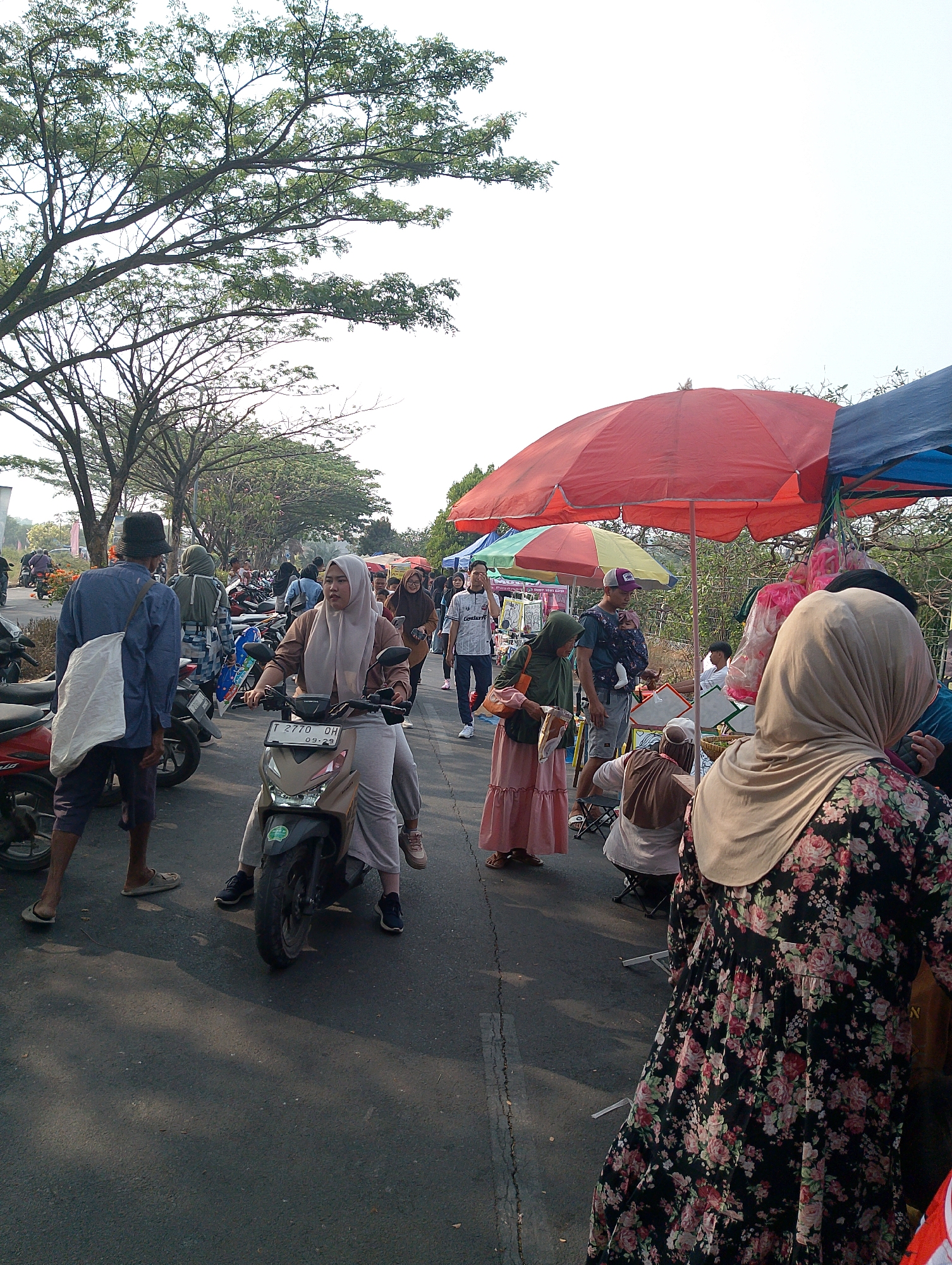 The image depicts a bustling outdoor market scene in Karawang, Indonesia, likely on a street or open area bordered by trees. The sky is bright, suggesting daytime. Several people are present, many wearing traditional Islamic clothing such as hijabs. In the foreground, a woman wearing a beige hijab and a brown top rides a scooter, with a license plate visible that reads "T 2770 DH 09-23". To her left, a person wearing a blue shirt and a black hat stands with a white bag slung over their shoulder, facing away from the viewer.
Further into the scene, various market stalls are set up, covered by large red and blue umbrellas. People are seen browsing, interacting, and walking. Motorbikes are parked along the sides of the road or path. The ground is asphalt, with some sunlight casting shadows. The overall atmosphere appears lively and active, indicative of a local market or fair.