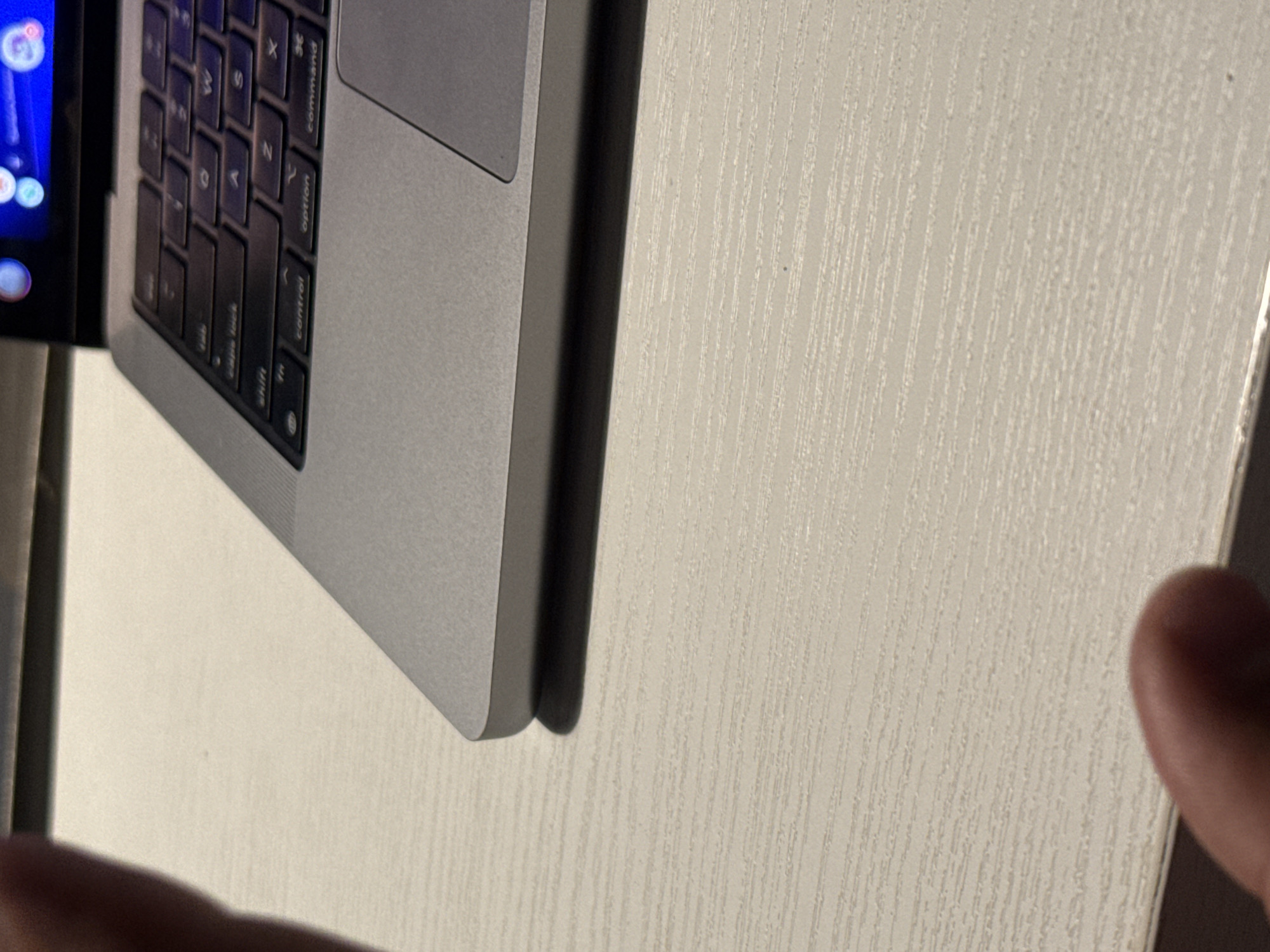 The image shows a close-up, angled view of a laptop keyboard and a textured surface, possibly a table. The laptop appears to be a MacBook, indicated by the layout and markings on the keys. Visible keys include "caps lock," "shift," "control," "option," and "command." The screen of the laptop is partially visible on the left, displaying colorful icons, suggesting it is powered on.
The textured surface to the right of the laptop has a wood-grain pattern with vertical lines. A blurred human finger is visible in the bottom right corner, partially obscuring the textured surface. The lighting suggests an indoor setting, possibly artificial light given the sharpness of the shadows and the bright screen.
No specific activity or event is clearly depicted, beyond the laptop being open and potentially in use. There are no discernible weather cues. The time of day is not evident. The location context provided is Kuta Selatan, Indonesia, but there are no visual cues within the image to confirm this specific location. The overall tone is neutral and focused on the details of the laptop and the surrounding surface.