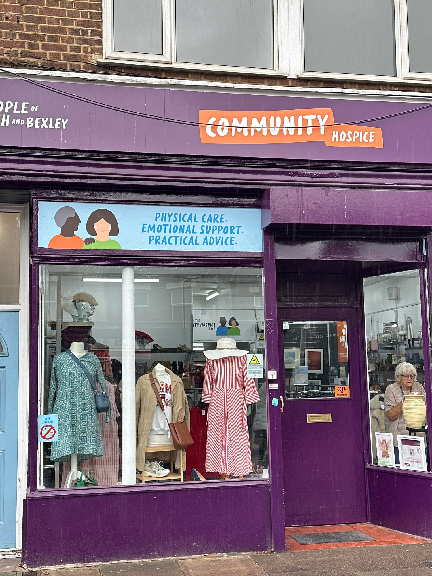 The image shows the exterior of a "COMMUNITY HOSPICE" shop in London, United Kingdom. The shop has a purple facade with a large display window. Inside the window, mannequins are dressed in clothing, and various items are displayed. A sign above the window reads "PEOPLE of BROMLEY and BEXLEY COMMUNITY HOSPICE" with an orange graphic. Another sign within the window displays an illustration of two people and the text "PHYSICAL CARE. EMOTIONAL SUPPORT. PRACTICAL ADVICE." To the right of the shop entrance, a woman is visible through the window, standing and looking at items on a shelf. The shop has a purple door and a doormat on the ground. The overall environment suggests a retail establishment in an urban setting.