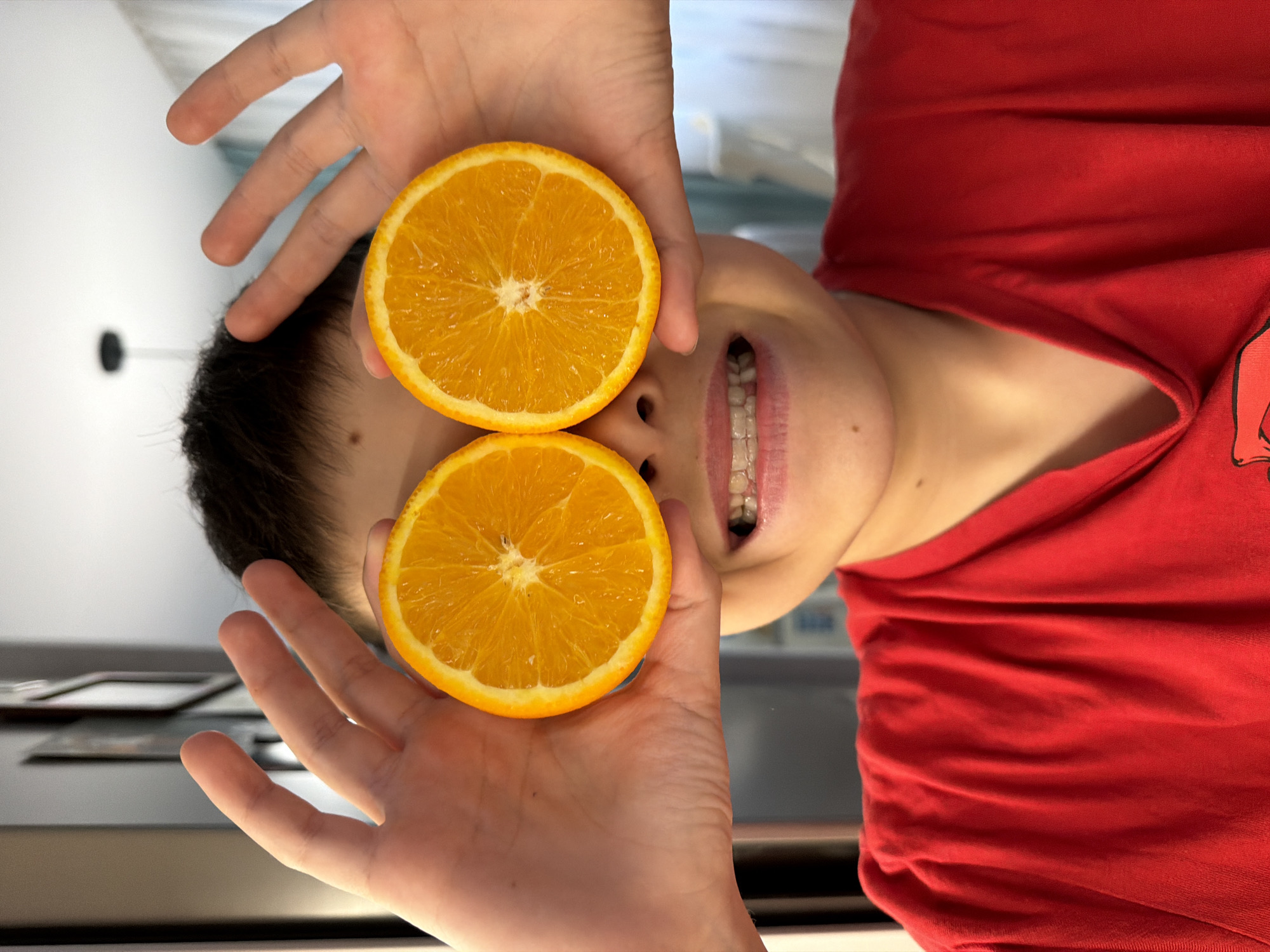 A person is holding two orange slices in front of their eyes, as if wearing orange slice glasses. The person has dark hair and is wearing a red shirt. The person is smiling and has braces. The background is a light-colored surface, possibly a countertop, with various objects in the background. There is nothing in the image to indicate a location in Votkinsk, Russia.
