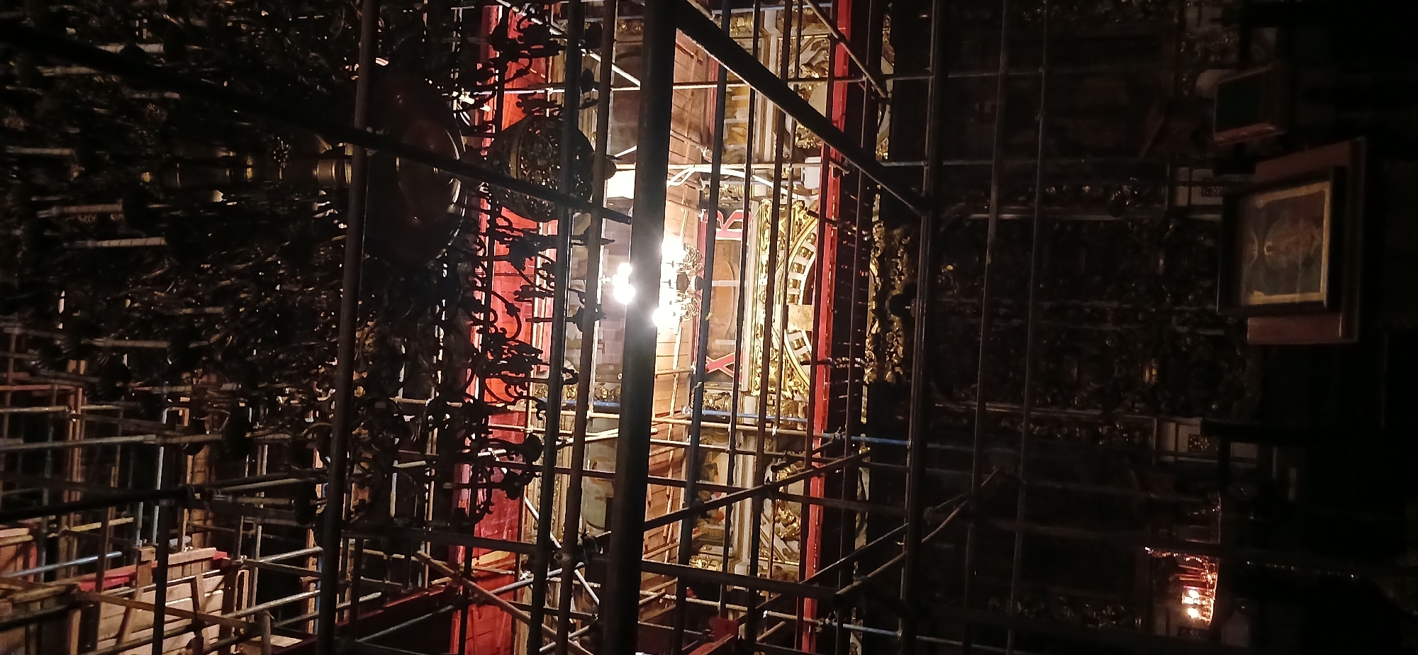 The image shows the interior of a building under construction or renovation, likely a historical or religious site, based on the presence of ornate golden details and scaffolding. A bright light is centrally located, possibly illuminating the work area. A picture frame can be seen at the edge of the image to the right. Scaffolding obstructs the view of the interior with its grid-like pattern. Given the prompt, the location of this building is likely Moscow, Russia. There are no visible people or actions taking place in the image. The overall environment is dark and cluttered with construction materials.