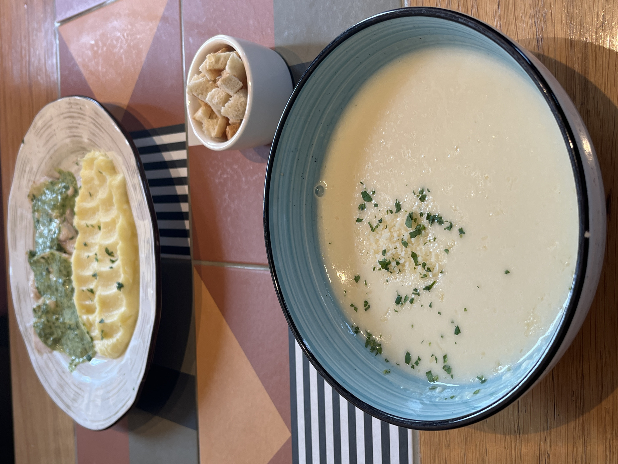 The image shows a top-down view of a table setting, likely in a restaurant. A blue-rimmed bowl of creamy soup, garnished with chopped herbs and possibly grated cheese, occupies the right side of the frame. Next to it is a small, white cup filled with croutons. On the left, there's an oval plate containing mashed potatoes and a green sauce or vegetable, possibly spinach or pesto. The table surface is decorated with a geometric pattern of brown, tan, and gray tiles, and there is a black and white striped object near the middle.
The location is claimed to be Verkhnyaya Pyshma, Russia, but there is no way to verify this from the image. The environment seems clean and well-lit, suggesting a pleasant dining atmosphere.