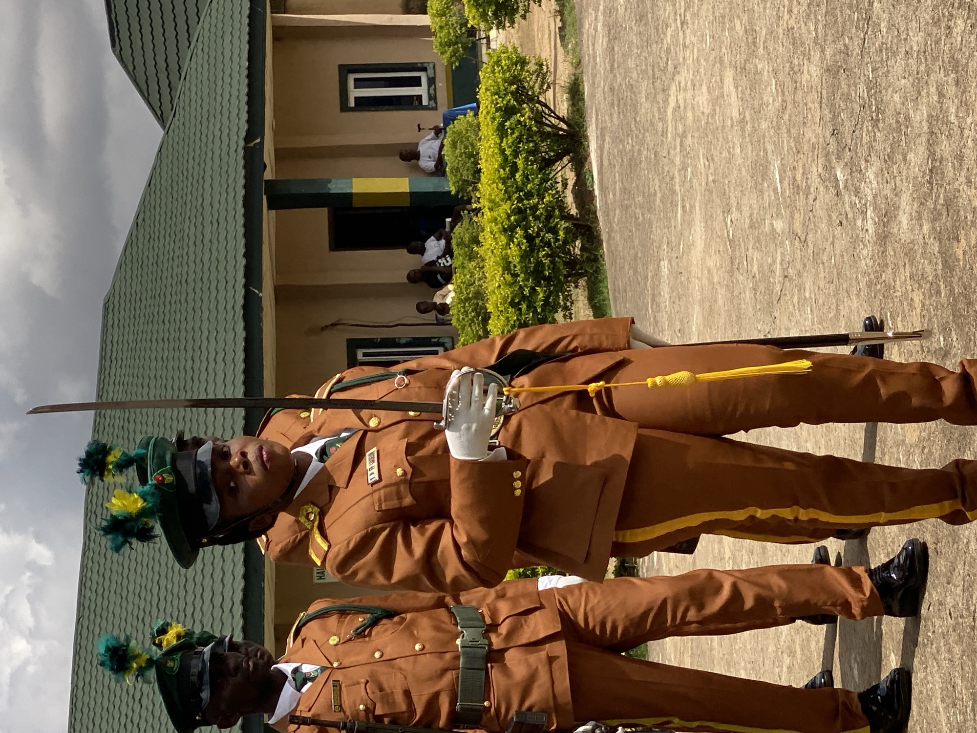 In Kaduna, Nigeria, a group of individuals in uniform stand on an outdoor paved area. The central figure, a woman, wears a brown uniform with gold trim, a green cap adorned with yellow and green feathers, white gloves, and holds a sword. To her left, a man in a similar brown uniform with a green cap stands at attention, holding a rifle. Two other individuals in brown uniforms are partially visible behind them. In the background, a building with a green roof and beige walls is visible, with several people seated and standing on a covered walkway. Lush green bushes line the front of the building. The sky is overcast.