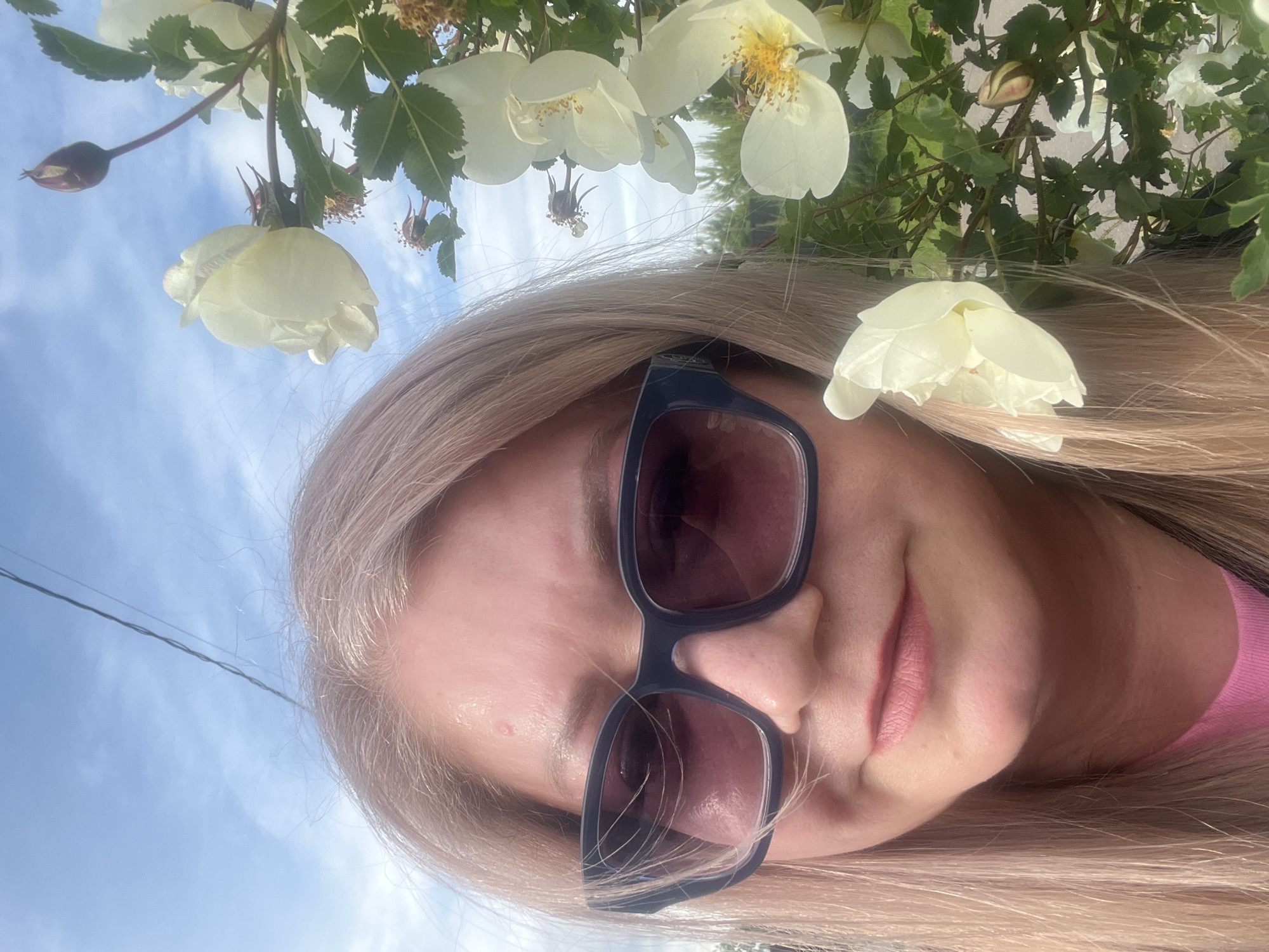 A selfie-style portrait features a blonde woman outdoors in Shud'ya, Russia, under a clear, bright blue sky with sparse white clouds. The image is rotated, showing the woman's head tilted to her right, with her long, light blonde hair flowing downwards and to her left. She wears dark-framed sunglasses that cover her eyes, reflecting a reddish tint. Her lips are slightly parted, revealing a subtle, relaxed expression, and she has light pink lipstick on. A pink garment is visible on her right shoulder and chest.
Overhead, from the viewer's perspective, are branches adorned with white or very pale yellow flowers, resembling wild roses, interspersed with green leaves. Some flowers are fully bloomed, showcasing yellow stamens, while others are still in bud form, and a few appear withered. These flowers create a natural, blooming canopy in the upper portion of the frame. In the top left, a dark, thin wire or power line stretches across the blue sky. The bright, natural lighting indicates it is daytime, likely morning or afternoon, with pleasant, sunny weather. There is no visible text in the image.