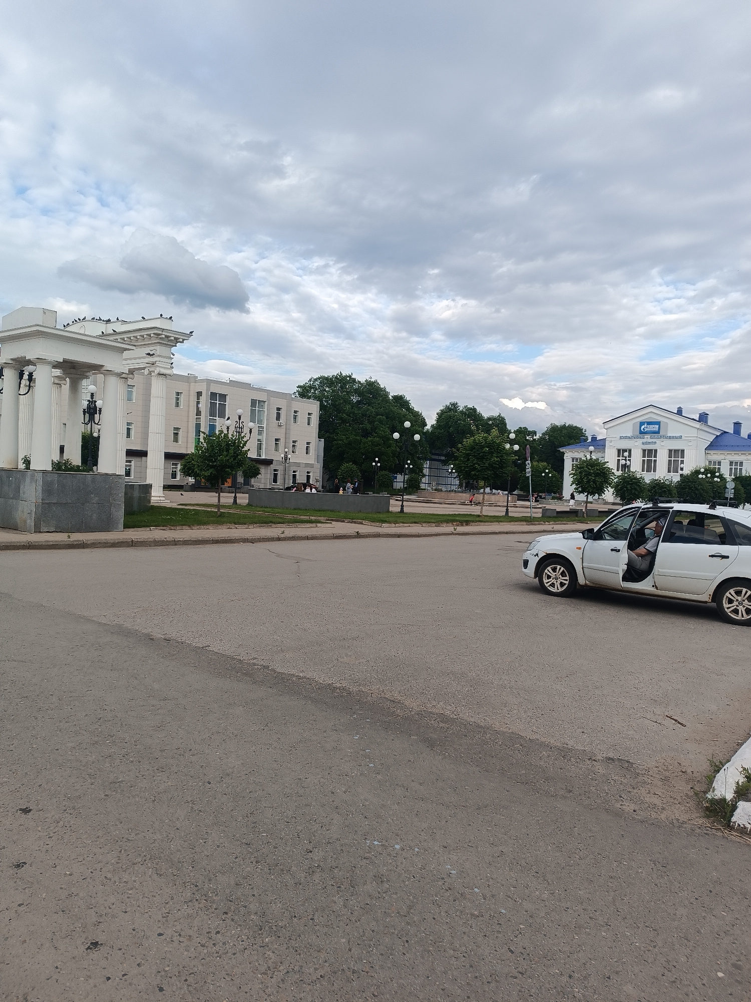 This is a full-frame, eye-level, medium shot of a street scene in Chaikovsky, Russia, under a cloudy sky. In the foreground, a white sedan is parked with its driver's side door open, and a person is visible in the driver's seat. The car is positioned on a paved road. In the mid-ground, there is a grassy area with trees and some people walking. To the left of the grassy area is a white building with columns, and to the right is another white building with a blue roof, featuring the "GAZPROM" logo and Russian text above the entrance. The overall impression is a typical urban street on an overcast day.