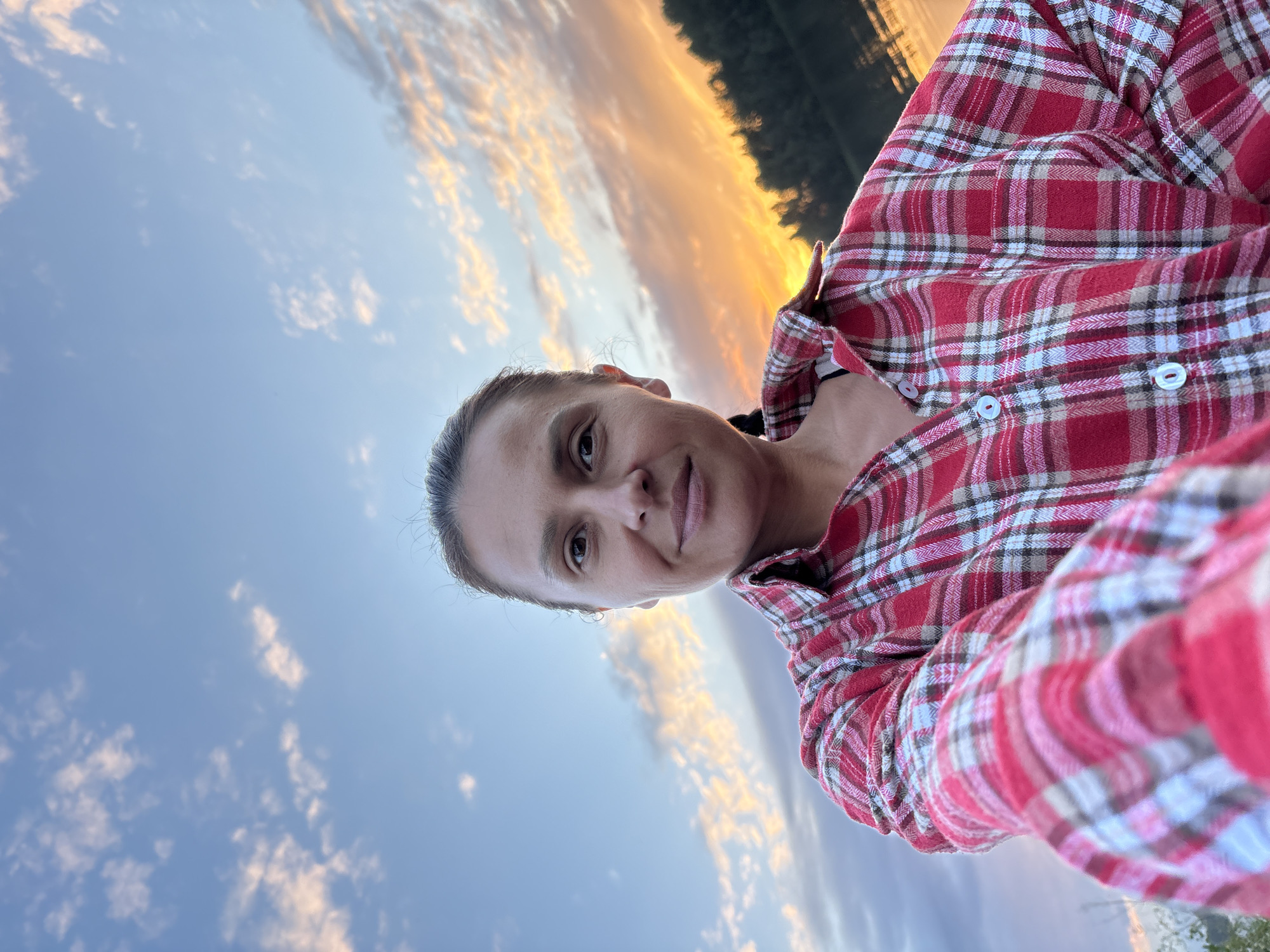 A middle-aged woman with her hair pulled back is taking a selfie outdoors. She is wearing a red and white plaid shirt. The background shows a sunset sky with clouds, a body of water, and a line of trees. The setting appears to be natural, possibly near a lake or river. The image quality is clear, and the lighting suggests it was taken during the golden hour. The location is identified as Chaikovsky, Russia.