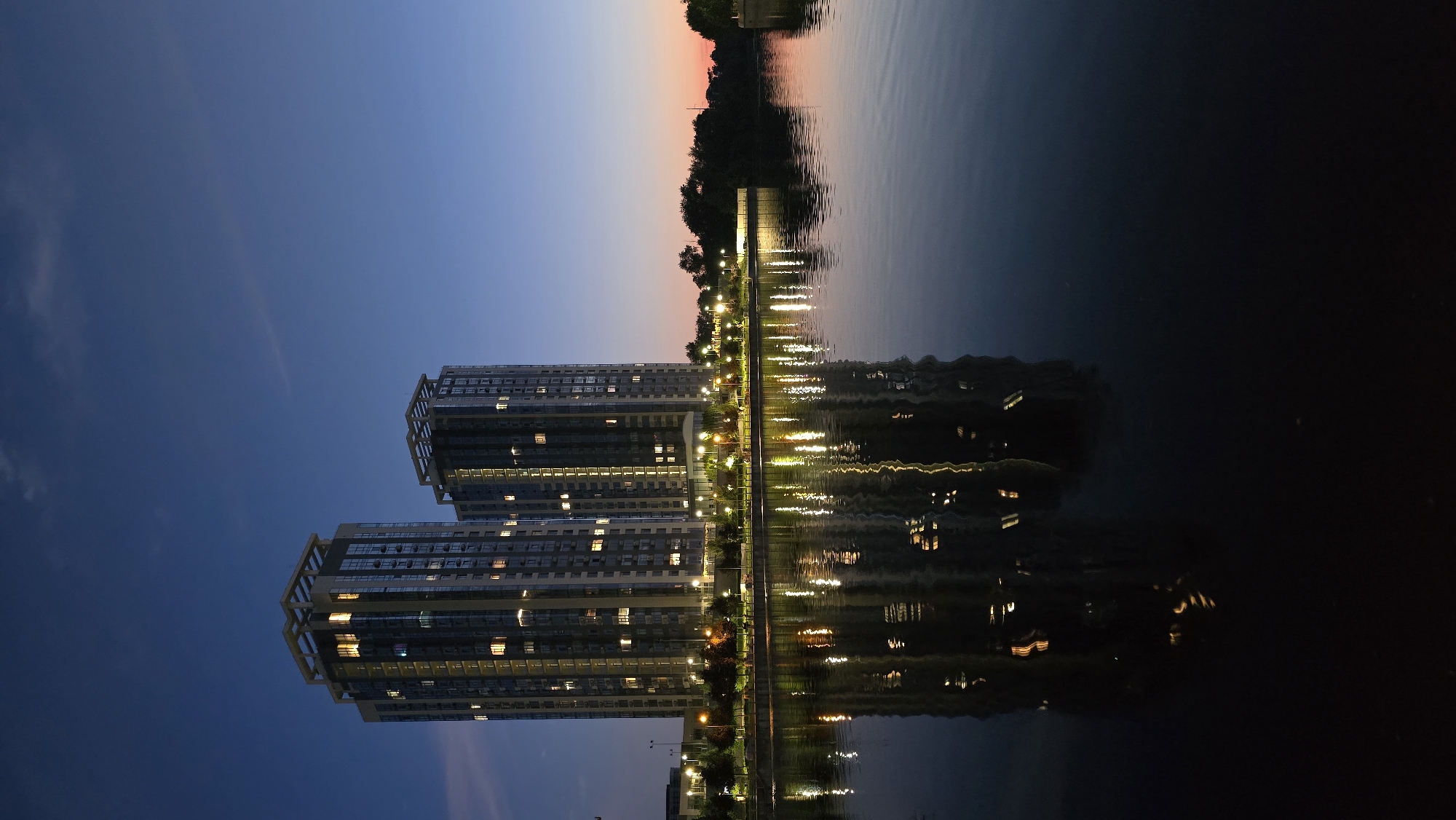This is a vertical, full-shot photograph taken at dusk, showcasing a modern residential complex with two tall buildings and their reflection in a calm body of water. The sky is a gradient of deep blue fading into a soft orange and pink near the horizon, suggesting the sun has recently set. The buildings, with their many windows illuminated by warm interior lights, stand prominently on the left side of the frame. A tree-lined promenade or park runs along the water's edge between the buildings and the water, with streetlights casting bright, yellow reflections on the water's surface. These reflections are distorted by slight ripples, adding a dynamic element to the otherwise still scene. The overall mood is serene and peaceful, with a touch of urban sophistication. There is no visible text in the image. The location context is Kyiv, Ukraine.