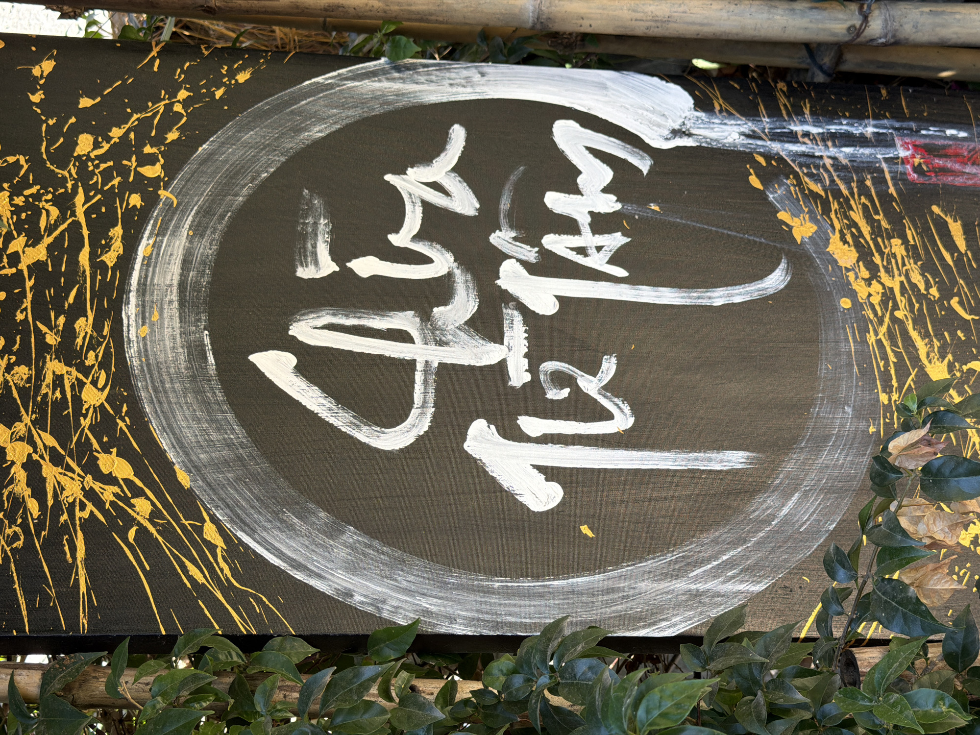 A sign in Đà Nẵng, Vietnam, features white calligraphy on a black background surrounded by a white circle. Gold paint splatters appear on the upper left corner and along the upper right edge. The sign is partially obscured by green foliage at the bottom and a bamboo structure at the top.