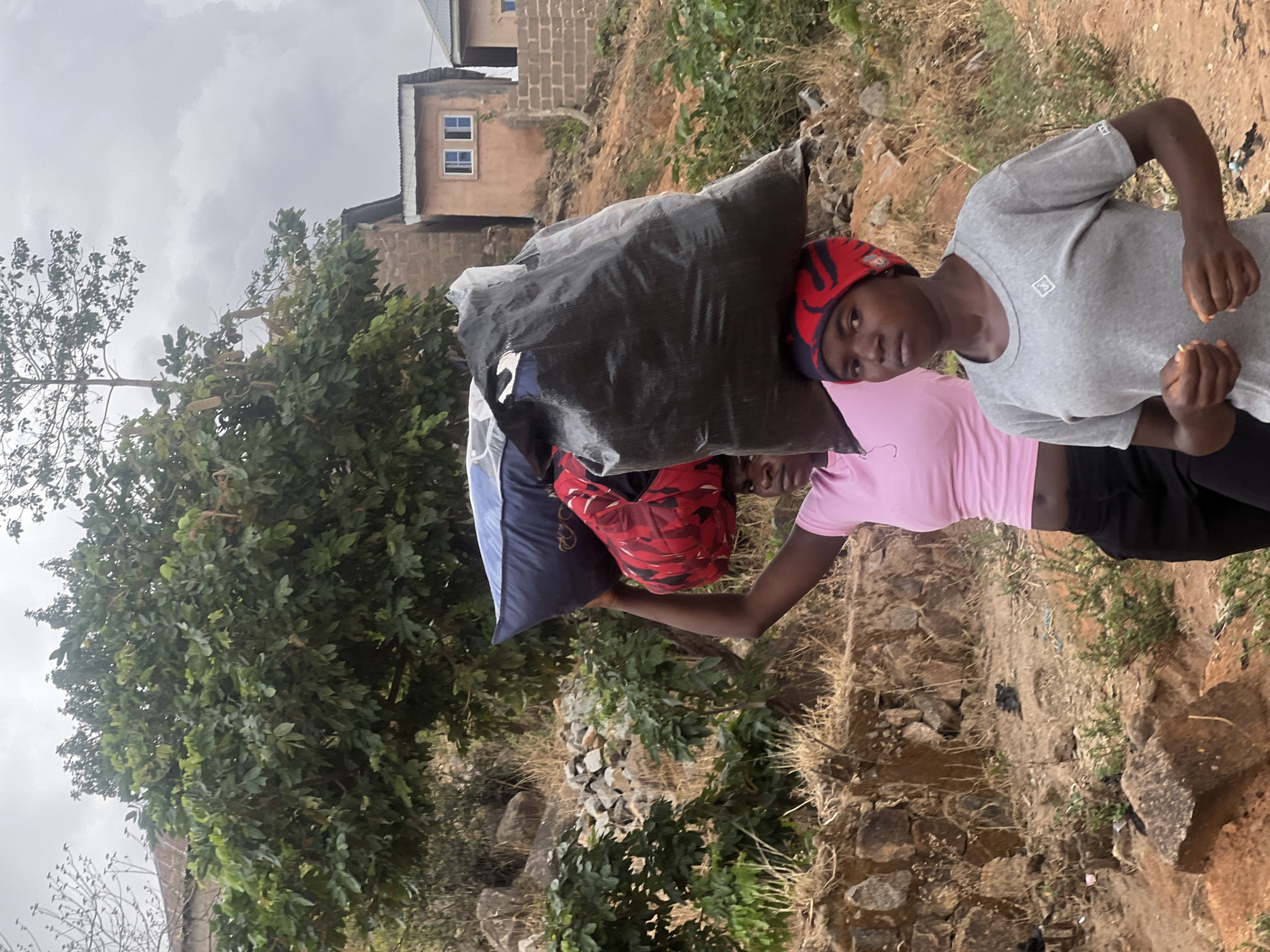 Two individuals stand on uneven terrain in Jos, Nigeria. One wears a grey t-shirt and a red patterned cap. The other wears a pink t-shirt and black pants. They are carrying large bags of unidentified materials on their heads. A building is visible in the background, along with trees and vegetation. The sky is overcast. The ground is rocky.