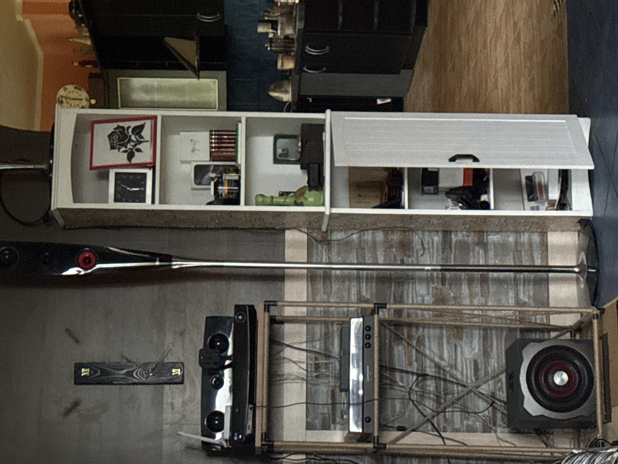 The image depicts an indoor scene, likely a living room or entertainment area, in what appears to be an apartment in Galinė, Lithuania. The overall atmosphere is cluttered with various electronic devices and decorative items.
On the left side of the frame, a tall, slender black floor speaker stands vertically. Next to it, a narrow, dark wooden clock with Roman numerals is mounted on a grey, textured wall. Below the clock, a black stereo system with multiple speakers is placed on a wooden shelf unit. Further down on the same shelf unit, a silver turntable is visible, with another black audio device stacked on top of it. At the bottom of this shelf unit, a black subwoofer with a red and silver center is positioned. The shelf unit itself is constructed of light-colored wood and black metal supports.
To the right of the speaker and shelf unit is a tall, white, open-shelved cabinet. The top shelf displays a framed black rose illustration next to a small black clock. Below this, various items are arranged, including what appears to be a vintage record player and books. The cabinet door is slightly ajar, revealing more items on a lower shelf.
In the background, a black kitchen cabinet unit with stainless steel handles is visible, suggesting the area may be adjacent to a kitchen. The floor is a mix of dark blue tiles and wooden-patterned flooring. The lighting in the room appears to be a mix of natural and artificial sources, creating shadows and highlighting different areas of the scene. There are no people present in the image.