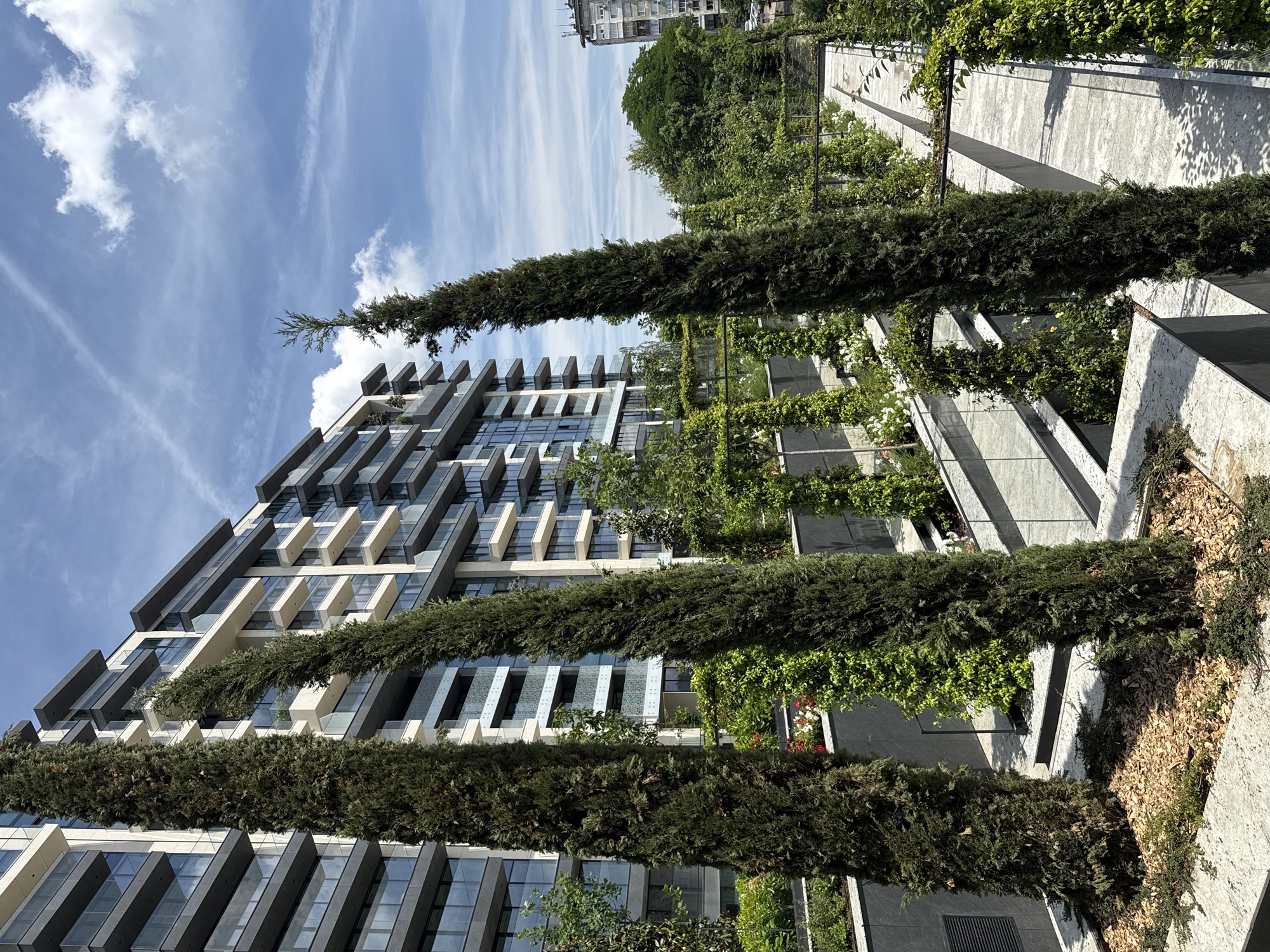 A modern, multi-story building, likely an apartment complex, stands prominently on the left side of the frame, characterized by its contemporary design featuring angular balconies, large reflective glass windows, and a facade combining light-colored and dark grey elements. In front of and extending beyond the building's height are several strikingly tall, slender evergreen trees, possibly cypress, arranged vertically.
To the right, a lush, multi-tiered landscape or terraced garden area unfolds. This green space incorporates planter boxes with textured light grey walls and darker bases, filled with diverse foliage including dense green vines climbing trellises and walls, as well as clusters of white and red flowers. The meticulous landscaping creates a visually appealing integration of nature within the urban setting.
The scene is captured on a bright, clear, and sunny day, indicated by the vivid blue sky dotted with scattered white clouds and the strong, distinct shadows cast by the trees and architectural elements. The overall atmosphere conveys a sense of modern tranquility and biophilic design. In the far background, behind the contemporary structures, glimpses of an older building and additional trees are visible. No people are present, and no specific activities are taking place. There is no visible text. The location context provided is Sofia, Bulgaria.
