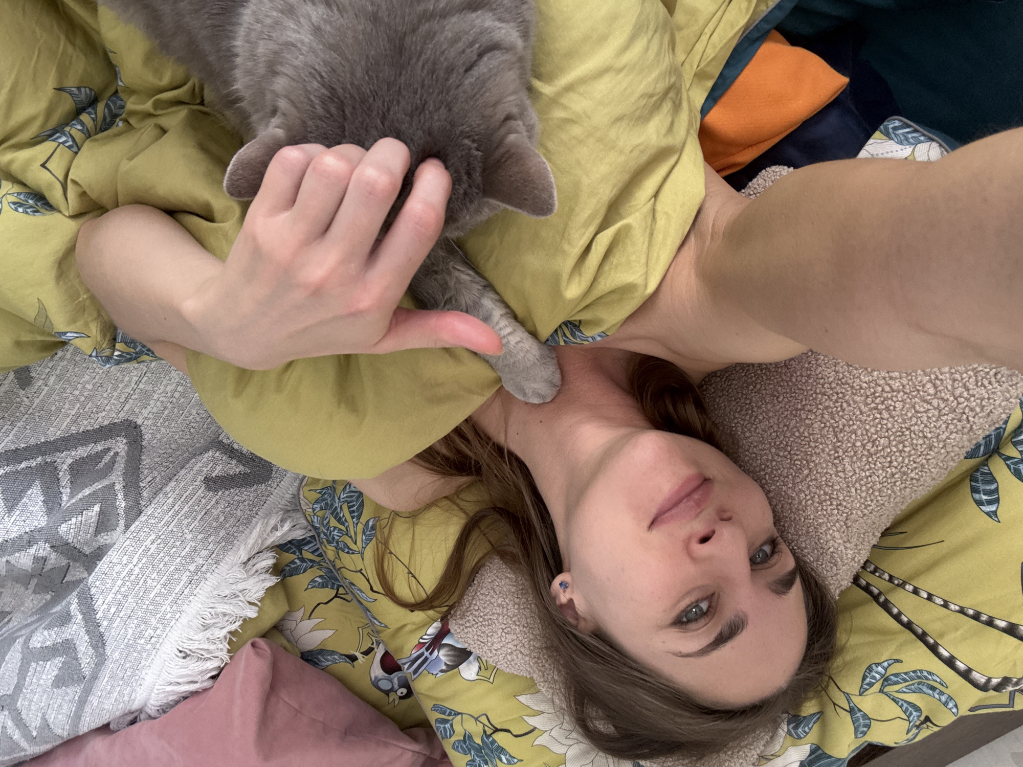 A woman lies on a bed with yellow and blue floral patterned sheets and pillows. She is interacting with a gray cat that is resting on her. The woman's right hand is on the cat's head, and the cat's paw rests on her neck. The woman has fair skin, brown hair, and blue eyes, and she is looking directly at the camera. There are various other pillows surrounding her, including orange, blue, pink, and tan colored pillows. The scene is indoors and appears to be a domestic setting. There is no information suggesting that the location is in Saint Petersburg, Russia.
