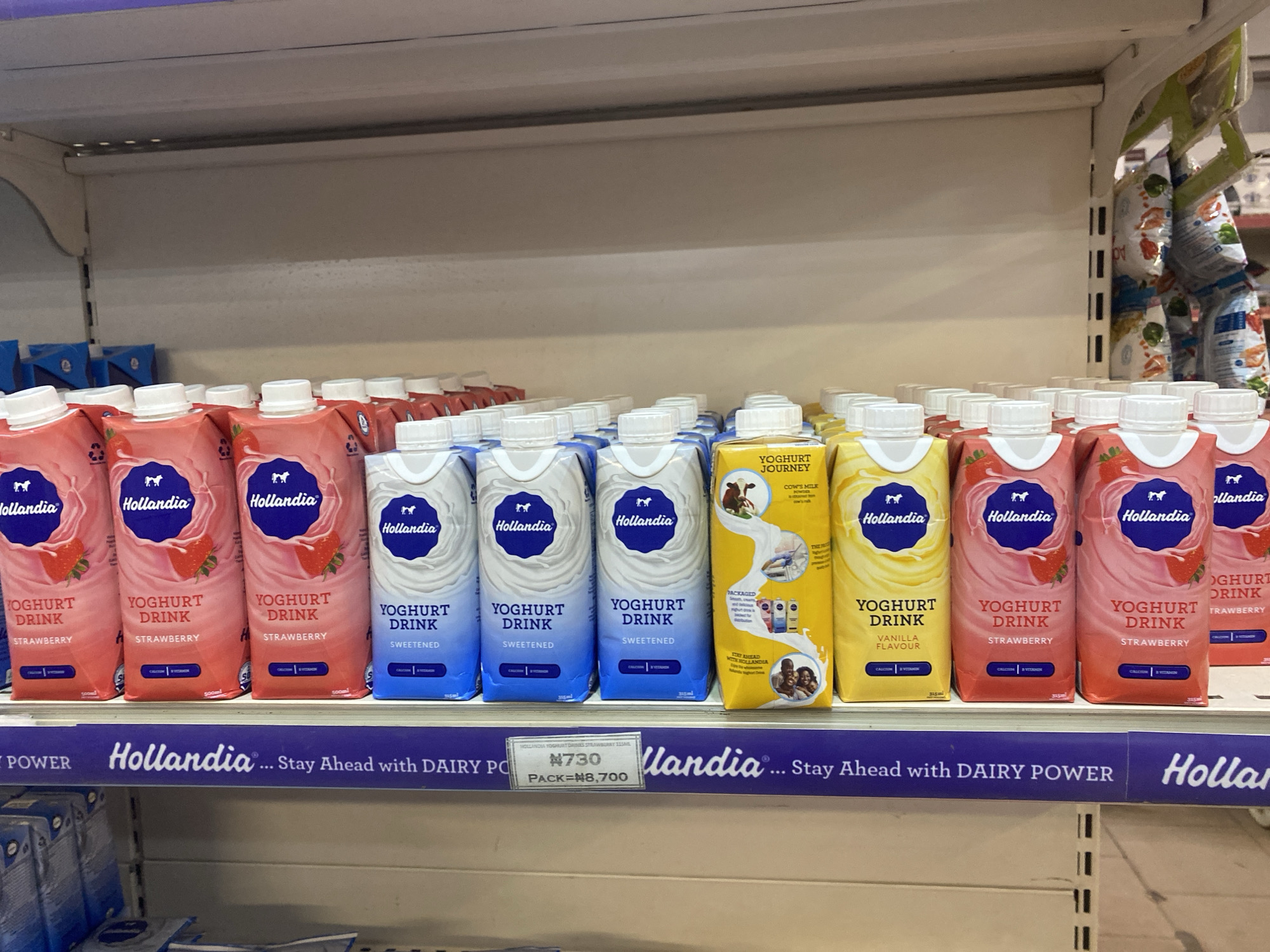 A retail store shelf displays rows of Hollandia yoghurt drink cartons in various flavors, including strawberry and vanilla. The cartons are primarily pink and blue, with a distinctive blue oval logo featuring a horse. Some cartons have images of strawberries. A price tag is visible on the shelf, reading "#730 PACK=#8,700". The shelf is illuminated by overhead store lighting, and the background consists of a neutral-colored wall. Based on the context provided (Hollandia products are common in Nigeria and the price format is consistent with Nigerian currency), this scene is likely from a retail establishment in Ikorodu, Nigeria, such as a supermarket or convenience store. There are no people visible in the image.