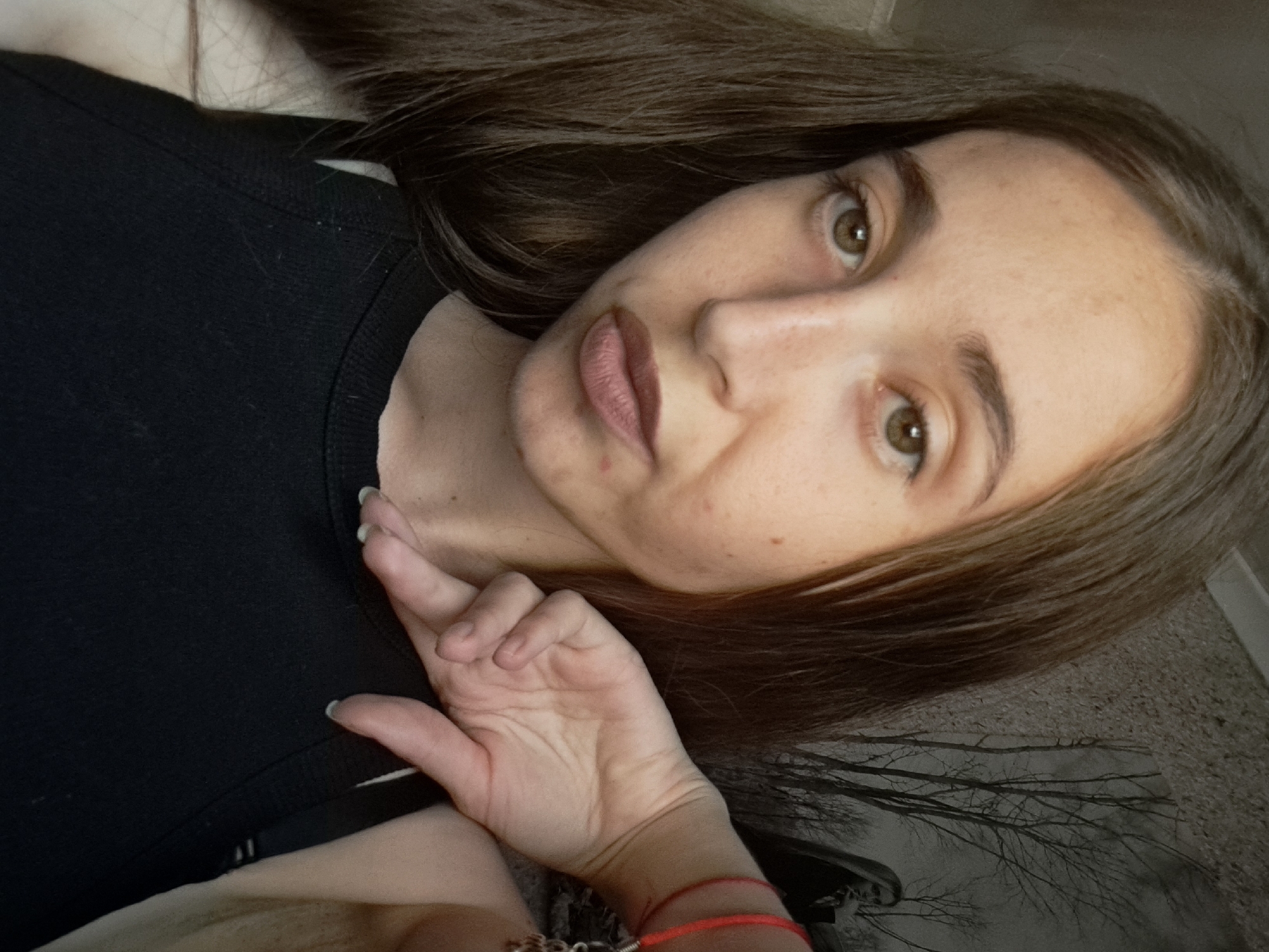 A selfie of a young woman is presented, taken indoors. The woman has long, brown hair that falls over her left shoulder and partially obscures her face. She has brown eyes and fair skin with some blemishes. She is wearing a black tank top. Her right hand is raised, with her fingers resting near her chin and collarbone, and a red bracelet is visible on her right wrist. In the background, a black and white image is visible on the wall, depicting bare trees and a distant figure. The setting appears to be a private residence. There is no information available about the location in the city of Vypasne, Ukraine.