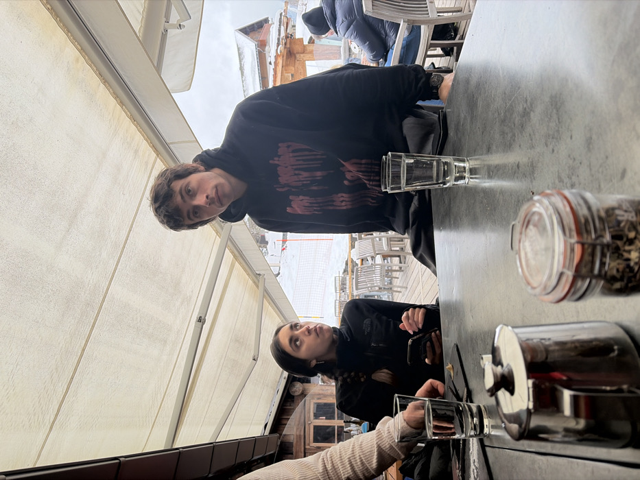 The image displays an outdoor dining or seating area under a light-colored retractable awning in La Clusaz, France, with a snowy mountain landscape visible in the background. Two young adults are seated at a dark, rectangular table in the foreground. A male, positioned right of center, wears a black hooded sweatshirt with a red graphic and a dark wristwatch on his left wrist; he looks directly at the camera. To his left, a female, with dark braided hair and a black zippered fleece jacket, holds a smartphone and looks upwards and to the left. The lower arm and hand of a third individual, wearing a light ribbed garment, are visible in the lower-left corner.
On the table are three clear drinking glasses, two of which contain water. Also present are a small metallic lidded pot and a glass jar with an orange seal containing dry, multicolored botanical material. Beyond the table, a wooden deck extends, furnished with numerous light-colored outdoor chairs; some are occupied by other individuals dressed in winter clothing. The background features extensive snow cover, ski slope markers (orange and yellow), and rustic wooden chalets with snow-covered roofs, all under an overcast sky.