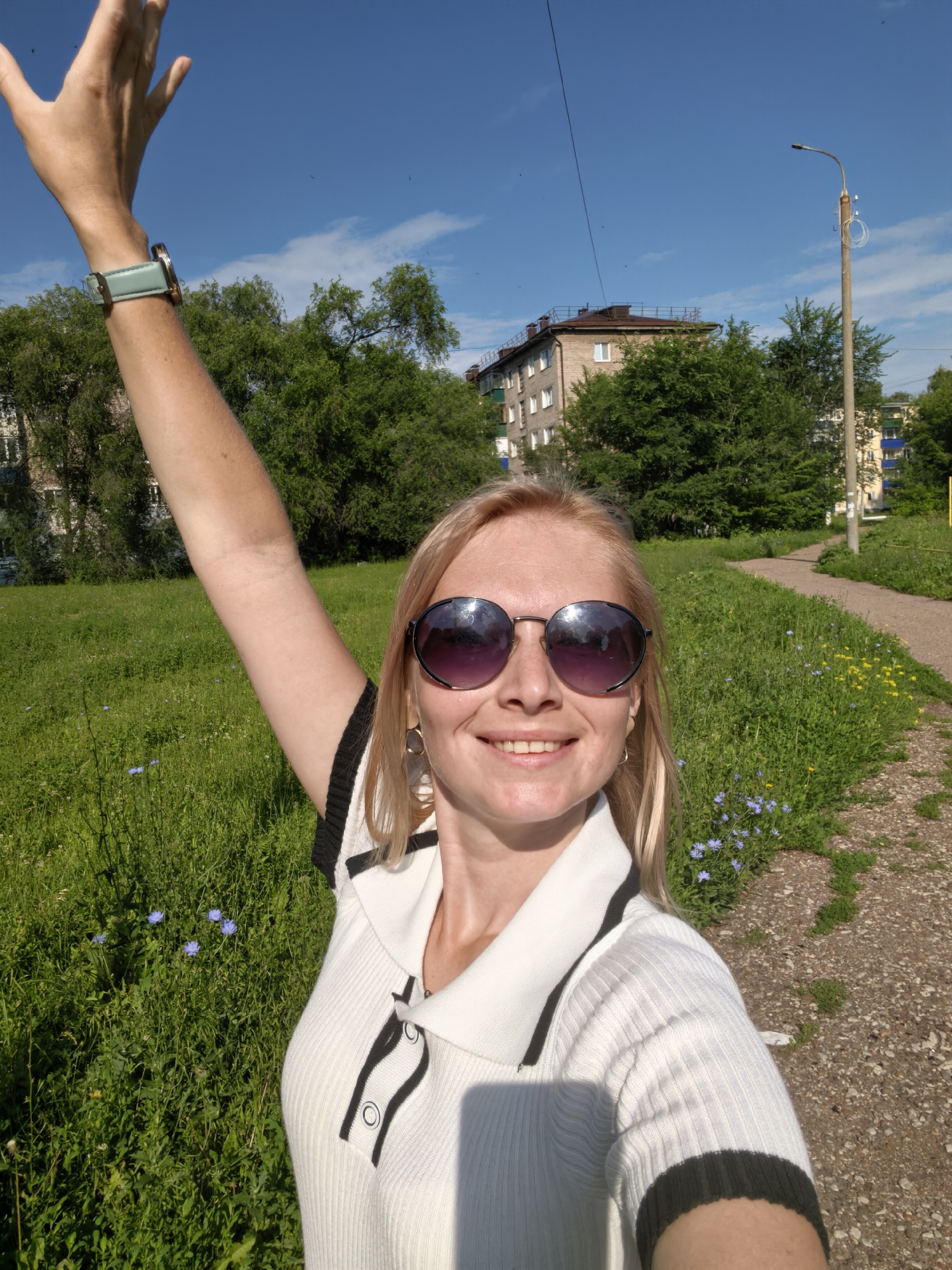 In a selfie taken outdoors on a sunny day, a smiling blonde woman with sunglasses and a white and black polo shirt is visible from the chest up. Her left arm is raised. In the background, a paved pathway winds through a grassy area with scattered blue and yellow wildflowers. Beyond the path, trees and multi-story apartment buildings are visible under a clear blue sky. The scene appears to be a residential area in the city of Sterlitamak, Russia.