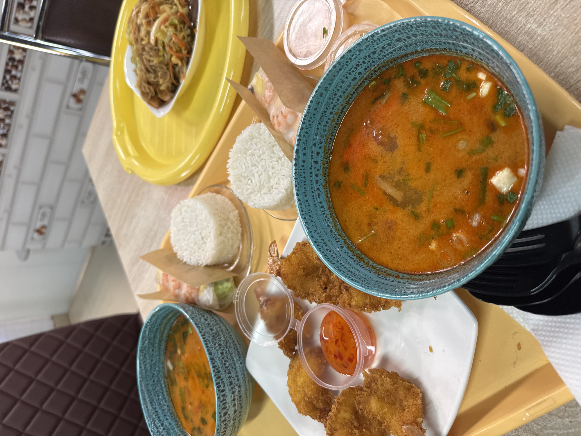 The image shows a close-up of a yellow tray laden with various Asian-inspired dishes. In the top left corner, there's a yellow oval dish containing a mix of noodles and vegetables. Adjacent to it, toward the right, are rice paper rolls partially wrapped in paper, along with a container of sauce. Two blue bowls filled with a reddish-orange soup are also visible. A white plate sits toward the lower-center of the frame, holding fried shrimp or similar items, accompanied by two small containers of reddish sauce. The tray appears to be placed on a light-colored table. The location is in the city of Unknown, Unknown.