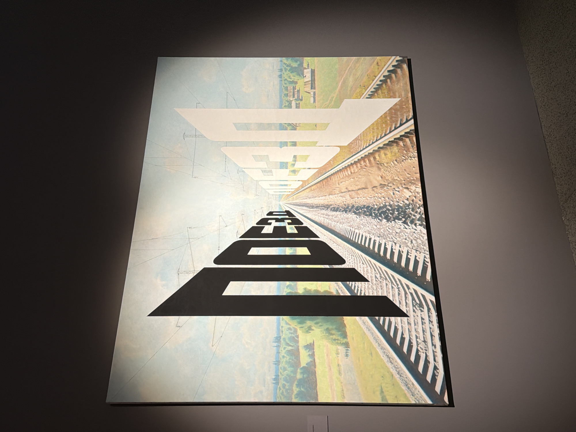 This is a vertical, eye-level photograph of a large painting hanging on a wall. The painting depicts a surreal landscape that combines elements of industrial infrastructure with nature. The foreground and middle ground are dominated by a set of railroad tracks that recede into the distance, creating a strong sense of perspective. The tracks are detailed, showing individual ties and gravel.
Overlaying the landscape, and seemingly emerging from or integrated with it, are large, stylized black and white letters. At the top of the painting, the letters are white and appear as if cut out from the sky, forming the word "BODEN". Below this, the letters are black and larger, forming the word "BODEN" in reverse perspective, as if they are on the ground and moving away from the viewer. The letters are stylized with sharp, geometric angles.
The background of the painting shows a hazy, pastoral landscape with fields, trees, and some distant structures that resemble rural buildings or possibly some industrial elements like chimneys or small towers. The sky is a pale blue with wispy clouds, and faint lines suggesting overhead wires or power lines are visible, further blending the natural and industrial themes. The overall mood is somewhat disorienting and thought-provoking due to the unusual juxtaposition of elements and the distorted perspective of the text. The lighting suggests an indoor setting, possibly an art gallery, with shadows cast on the wall to the left of the painting. There is no discernible time of day or weather indicated within the painting itself, beyond the depicted sky. The painting is the sole focal subject.