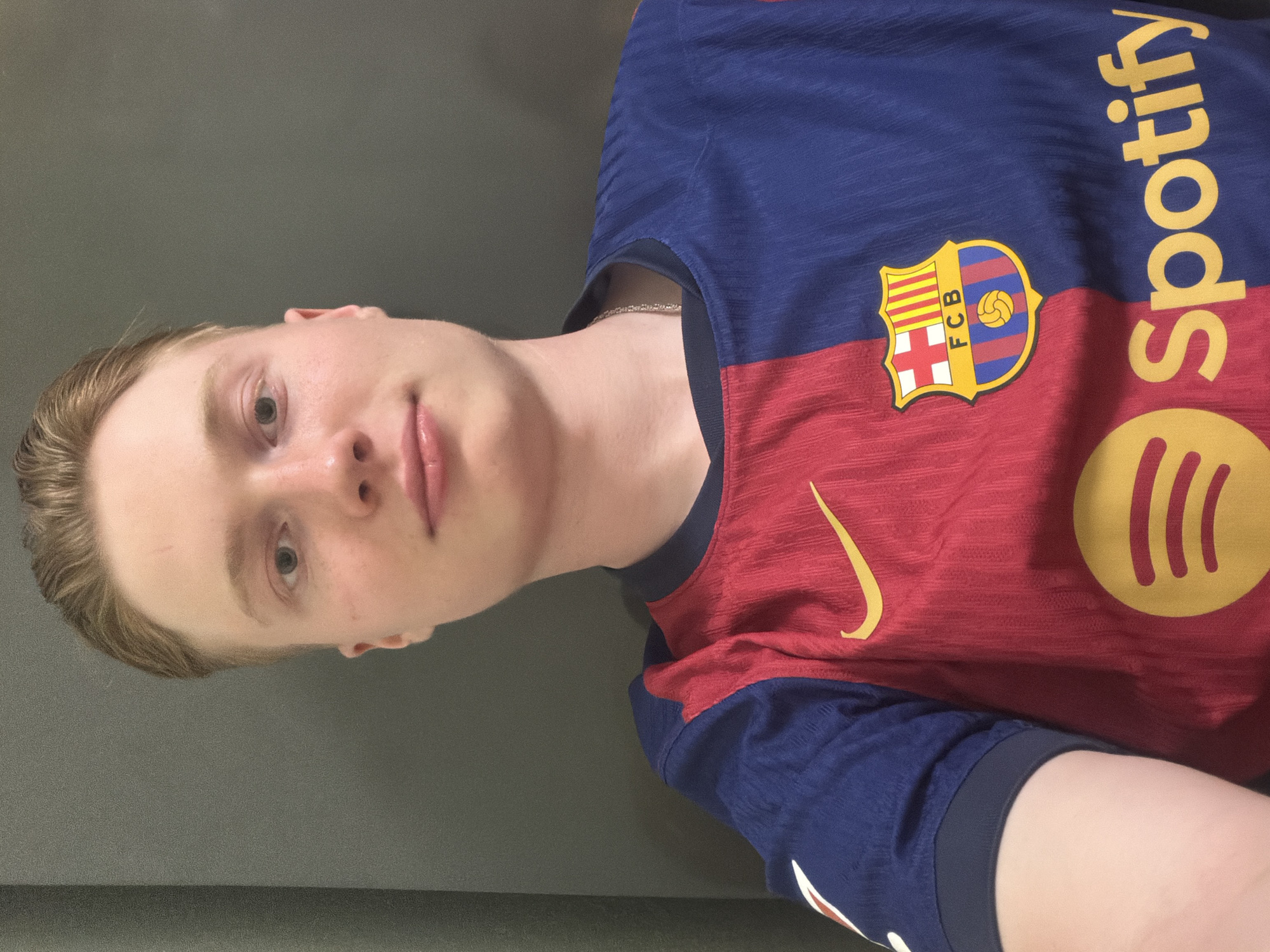 A young man with fair skin and light brown hair is pictured in a close-up, waist-up selfie. He is wearing a blue and red vertically split soccer jersey with a Nike logo on the red side and the FC Barcelona crest and Spotify logo on the chest. The background is a solid gray surface, appearing to be a wall or furniture. The lighting is soft and even, illuminating his face from the front. He is looking directly at the camera with a neutral expression.
