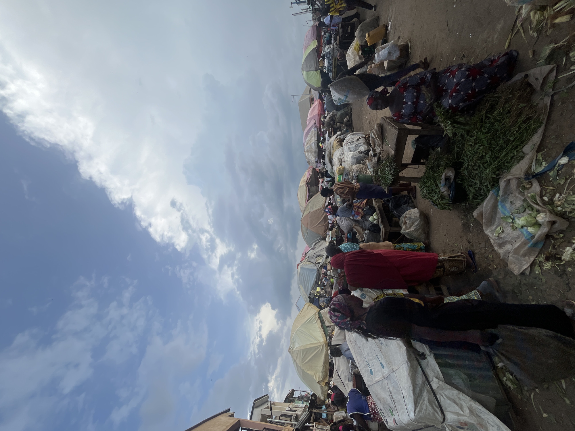 The image depicts a bustling outdoor market scene in Rafin Par, Nigeria. The foreground and midground are filled with people, many of whom are likely vendors and shoppers. They are dressed in a variety of traditional and modern clothing. Various stalls and displays are visible, featuring goods such as vegetables, produce, and other market items. Umbrellas are strategically placed over some of the stalls, suggesting a need for shade from the sun or protection from potential rain. Large sacks and bundles of goods are stacked, indicating the commercial activity taking place. The ground appears to be dirt or unpaved. The sky is partially cloudy, with patches of blue sky visible, suggesting it could be daytime. The overall atmosphere appears to be one of lively commerce and daily life within the community.