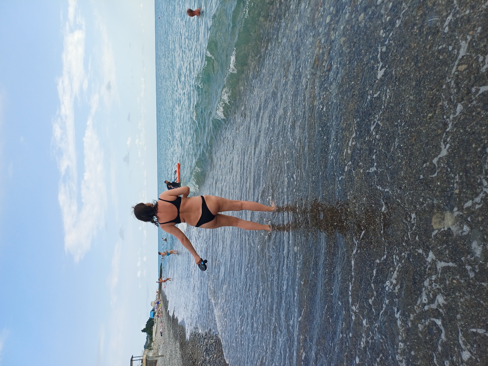 The image shows a beach scene in Sochi, Russia. The primary subject is a woman in a black bikini standing in shallow water at the edge of the sea, with her back to the camera. She is holding black flip-flops in her right hand. The water is clear with small waves and foamy edges, lapping over a pebble beach. In the background, other people are visible in the water and on the shore. The sky is blue with scattered white clouds, suggesting it is daytime with pleasant weather. The sea is calm and blue, stretching to the horizon. The overall atmosphere appears to be relaxed and summery. There is no visible text in the image.