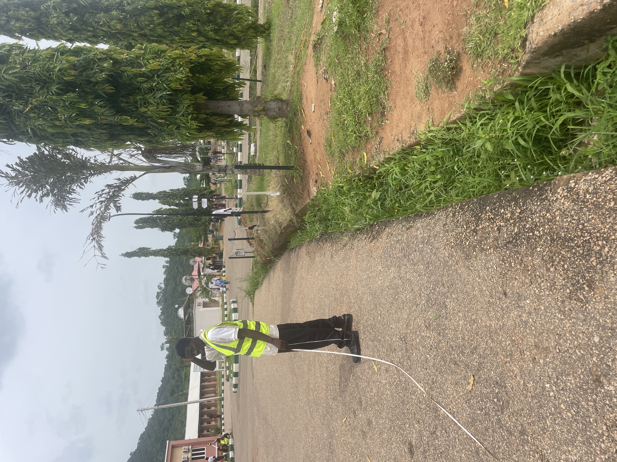 A person in a high-visibility yellow vest and black pants stands on a paved path. The person is facing away from the camera, looking down at a white cable that stretches out on the ground. The cable appears to be a measuring tape or similar tool. In the background, there are trees with lush green foliage, a dirt path with sparse grass, and some buildings. The sky is overcast, suggesting a cloudy day. In the distance, a hillside covered in trees can be seen. Several other people are visible in the background, some walking and some standing near the buildings. The scene appears to be an outdoor campus or park area. The image is oriented vertically. The overall atmosphere seems to be that of an outdoor work activity or measurement being conducted.