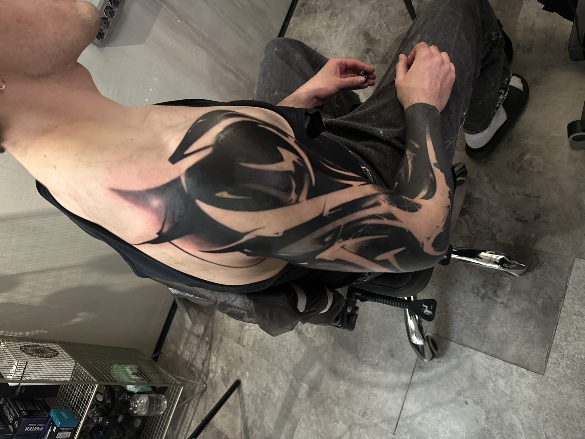 A high-angle shot depicts a person seated indoors in an office-style swivel chair. The person, a male, is seen from the upper torso, wearing a black tank top or undershirt and dark pants. A prominent black tattoo covers their left shoulder, upper arm, and extends onto the side of their torso. The tattoo features an abstract, tribal-like design with solid black areas and lighter, possibly negative space or shaded sections. The skin around the tattooed area appears slightly reddened, suggesting recent work or healing. The person's left hand rests on their lap, holding a small dark object. Their feet are visible, wearing black flip-flops with white socks.
The setting appears to be a modern indoor space with a light grey tiled or concrete floor and light grey walls. To the left of the frame, a metal wire basket holds various items, including a clear plastic water bottle and several boxes. Visible text on a black box clearly reads "ПЕРЧАТКИ" (Gloves) and "ЧЕРНЫЕ" (Black). Part of the text "VISAT" or "VISIT" is visible on a blue box. An electrical multi-plug outlet is mounted on the wall above the basket. The chair features chrome legs and a visible lever marked "LIFT". The lighting is bright and even, suggesting daytime or well-lit artificial conditions.