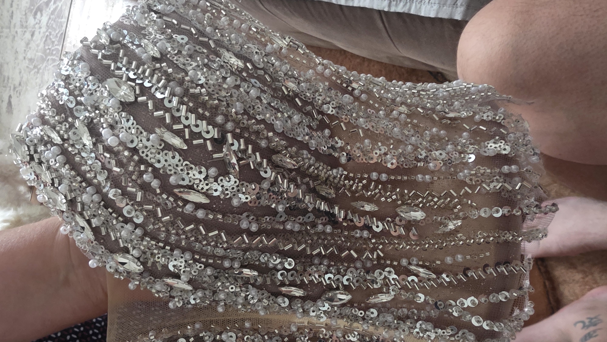 The image shows a close-up of a beige or light brown fabric intricately adorned with silver sequins, beads, and rhinestones. The embellishments are arranged in rows or patterns across the fabric. Parts of two human legs, including a knee, are visible in the upper right corner, and a portion of a hand is visible on the left. The fabric appears to be draped or lying on a surface with a light brown texture. The location is not discernible from the given image.