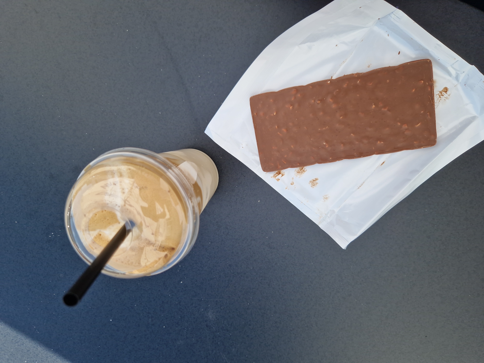 An overhead shot shows a clear plastic cup containing a frothy, light brown beverage with a black straw, placed on a dark, textured surface. To the right of the cup, a rectangular chocolate bar with visible nut pieces is unwrapped and resting on a white paper wrapper. The scene is illuminated, casting a shadow from the left side of the frame. The context suggests this image was taken at an outdoor cafe in Sofia, Bulgaria, likely during daylight hours.