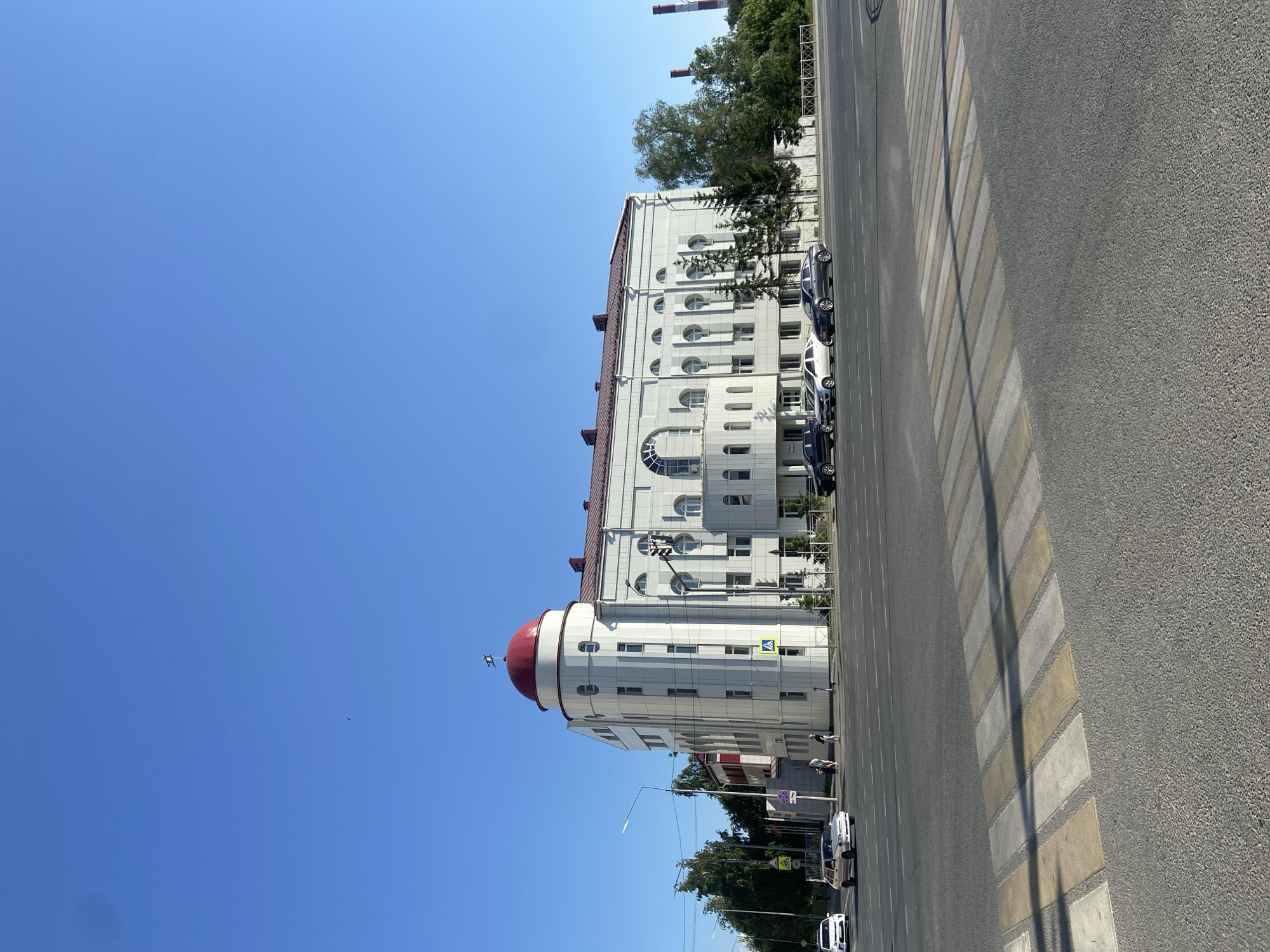 The image shows a street scene in Kazan, Russia. A white, multi-story building with a red domed corner structure dominates the left side of the frame. The building's architecture includes arched windows and a red roof trim. A crosswalk stretches across the foreground street, with parked cars lining the street in front of the building. The sky is clear and blue. There are some people walking by the building and on the sidewalk. Trees are visible in the background, along with smokestacks.