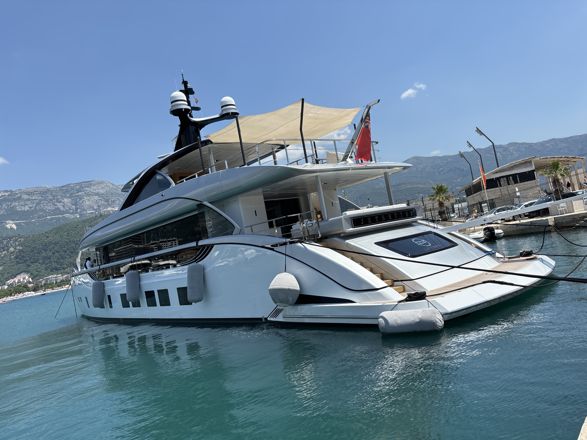 A white luxury yacht named "JETSETTER LONDON" is moored in the marina of Budva, Montenegro. The yacht has multiple levels, a light-colored awning, and is docked alongside other vessels and a promenade. The water is a clear turquoise. In the background, there are green mountains and buildings along the coastline. The sky is blue with a few white clouds. There are no people clearly visible on the yacht or in the immediate foreground.
