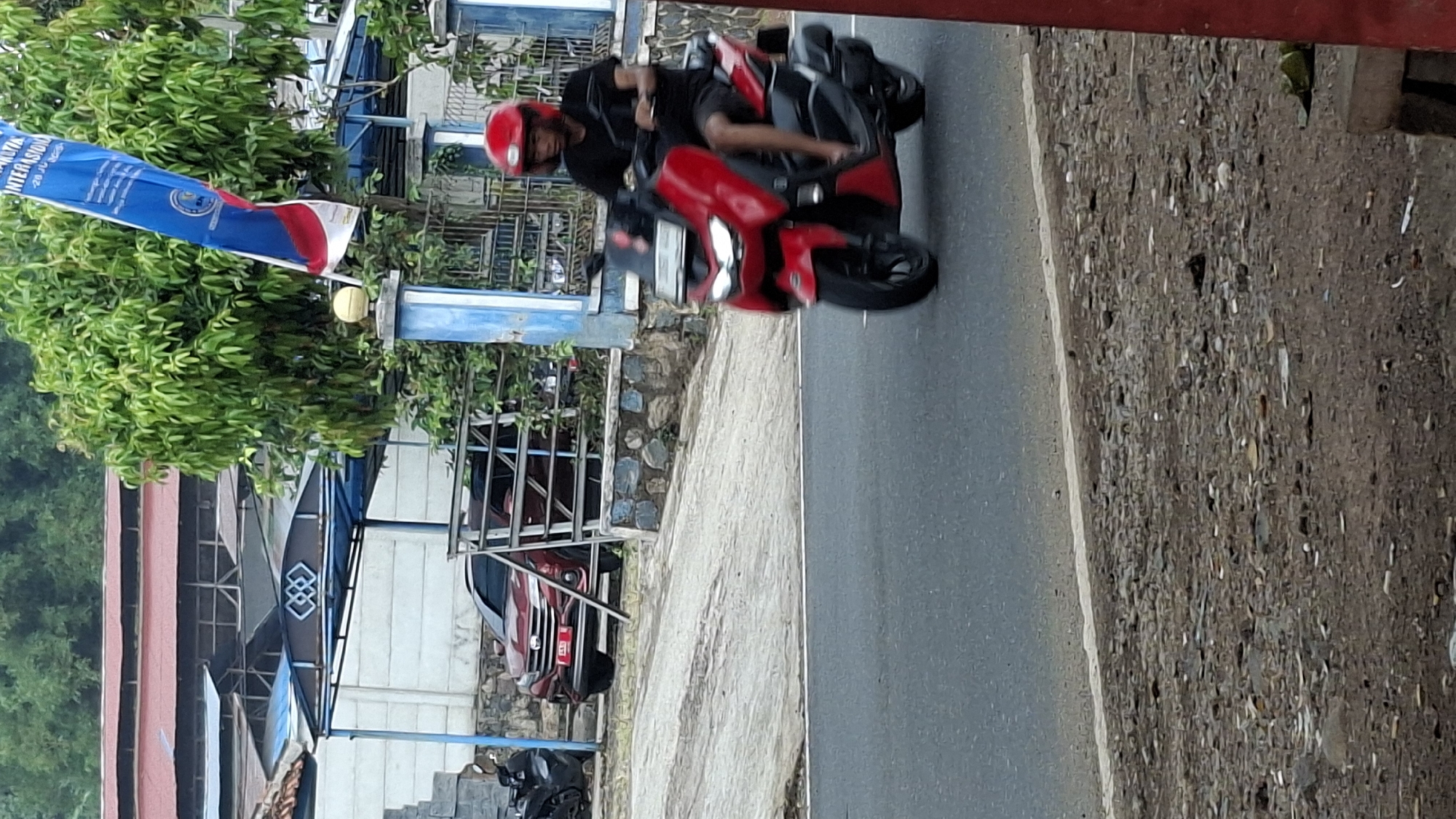 A man wearing a red helmet and black t-shirt rides a red motorcycle on a paved road in Cilaku, Indonesia. The man is facing forward and appears to be looking at the road. The motorcycle is red with black accents. The background shows trees, buildings, and a blue and white flag. The road is asphalt and appears to be in good condition. There are some scattered rocks and debris on the side of the road. The sky is overcast.