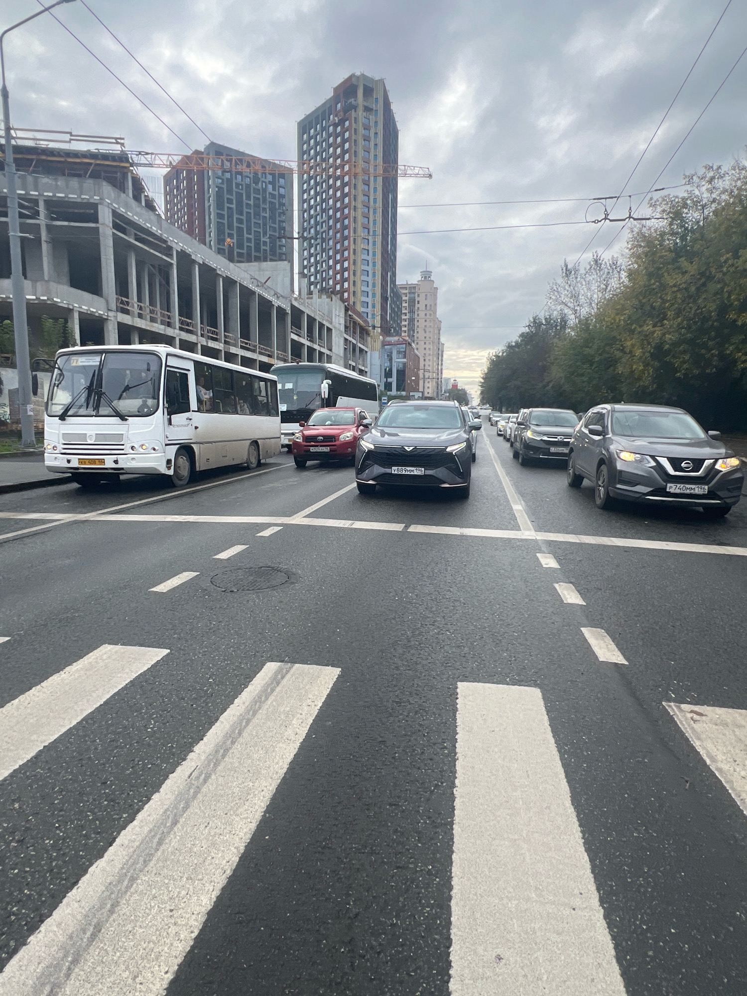 A grey, overcast day frames a street scene in Yekaterinburg, Russia, with a pedestrian crossing in the foreground and traffic at a standstill. A white bus, license plate "CXB 408 196," is positioned on the left side of the road, facing forward. To its right, a red SUV, license plate "M929 AK 196," is also stopped. Further down the road, a dark SUV, license plate "Y889MM 196," is visible, followed by a line of other vehicles. In the background, construction is underway on a multi-story building, with cranes and scaffolding present. Several other tall buildings, including residential towers, line the street. The environment appears urban, with trees lining the right side of the road.