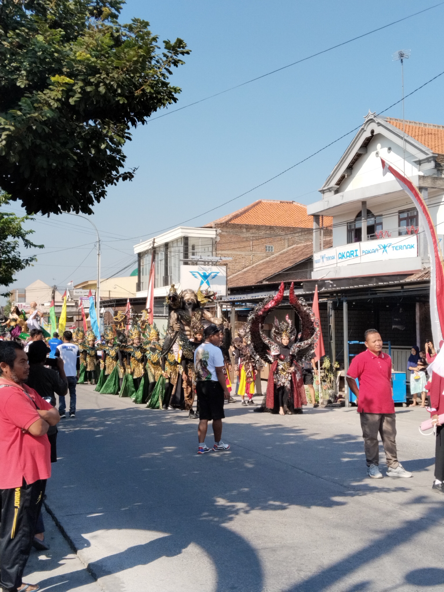 An outdoor daytime street parade is taking place on a sunny day in the city of Gondang, Indonesia. A line of people in elaborate costumes, some with large headdresses and ornate clothing in gold, green, and red, are walking down the paved street. Some individuals are dressed as mythical creatures or characters with detailed makeup and accessories. Several flags, including Indonesian flags (red and white), are being held up. Spectators are positioned on the side of the street, observing the procession. Buildings with red-tiled roofs and a white building with the signage "AKARI PAKAN TERNAK" are visible in the background. A large tree with green foliage is on the left side of the frame. The street is lined with shadows from trees and overhead power lines.