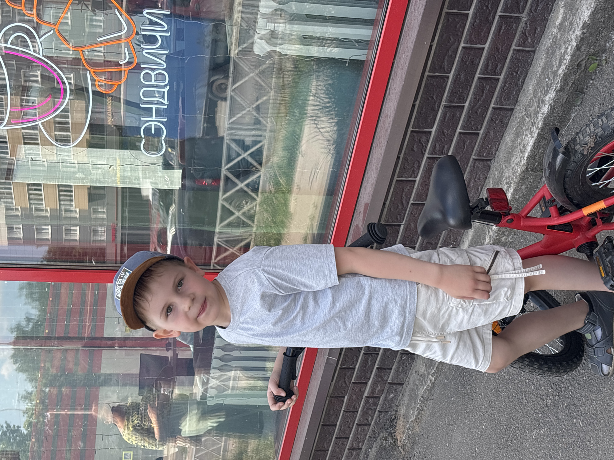 A young boy, appearing to be around five to seven years old, stands next to a red bicycle on a sidewalk in front of a building with a red-trimmed window. The boy is wearing a grey t-shirt, light-colored shorts, a blue and brown baseball cap, and grey sandals. He is looking towards the camera with a slight smile. Behind him, the window of the building reflects the exterior, showing a street with buildings and trees. A neon sign with Cyrillic script and images of a coffee cup and croissants is visible in the reflection. The building's facade is made of dark brown bricks. The pavement is grey asphalt. The overall scene suggests a daytime setting in an urban environment, likely a commercial area. The location is identified as Yaroslavl, Russia.