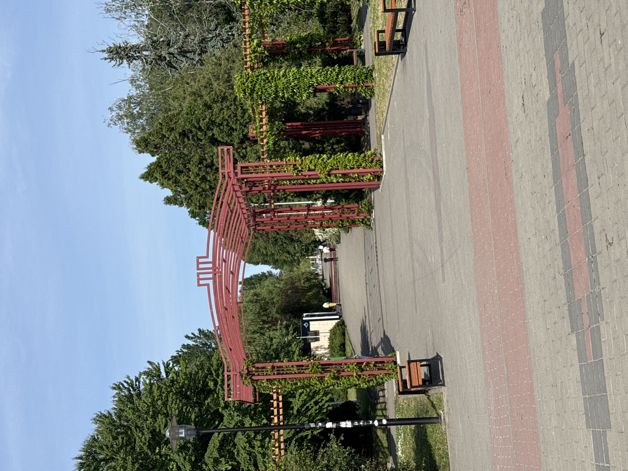 The image shows a pathway in a park, possibly in the city of Ternopil, Ukraine. A series of red metal arches create a tunnel-like effect, with greenery growing on and around them. The pathway is paved, with a brick pattern along the edge. There are benches near the pathway. The sky is blue.
