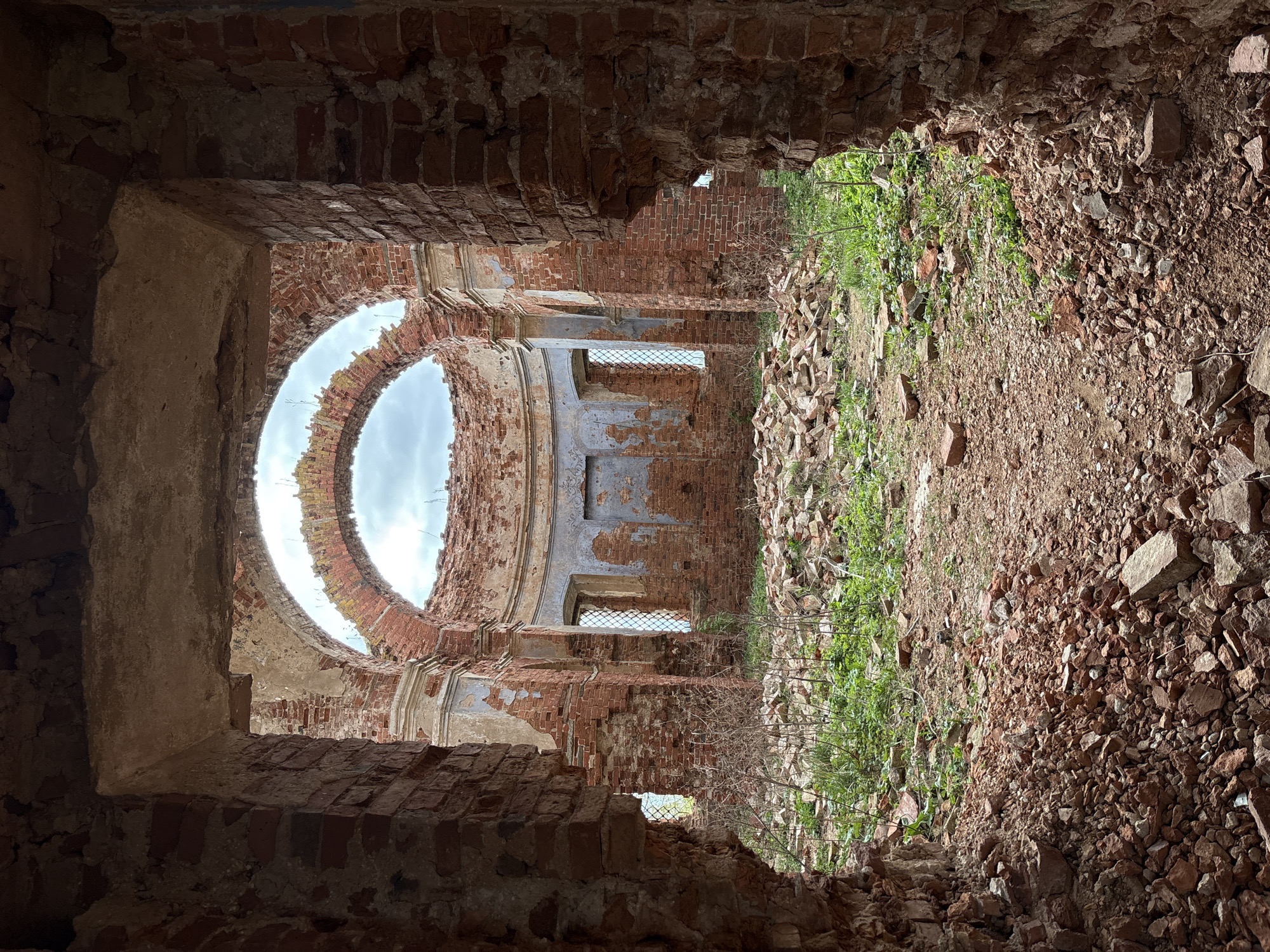 The image presents the interior of a severely ruined brick building, likely an abandoned church or a large historical structure in Staroe Rakomo, Russia. The photograph is taken from within a dark, narrow archway, looking into a larger, dilapidated space.
The most striking feature is a massive, partially collapsed circular opening in what appears to be the former dome or ceiling, through which a bright, overcast sky is visible. Below this, the curved interior walls are constructed from reddish-brown brick, with much of the original plaster and decorative finishes having eroded or fallen away, revealing the rough brickwork beneath. Traces of architectural elements, such as cornices, moldings, and pilasters, are still discernible on the crumbling surfaces. Several rectangular window openings are set into these walls; one retains what looks like a metal grate, while others are open to the outside.
The ground within the ruin is uneven and covered with a thick layer of debris, including broken bricks, stones, and earth, suggesting a long period of decay. Patches of vibrant green weeds and other vegetation grow vigorously amidst the rubble, particularly on the right side of the frame, indicating nature's reclamation of the abandoned space. The lighting is natural, with diffused light from the overcast sky illuminating the interior, while the foreground and the entry archway are cast in deeper shadow. There are no people visible in the scene, and no discernible text. The image evokes a sense of decay, history, and the relentless passage of time.