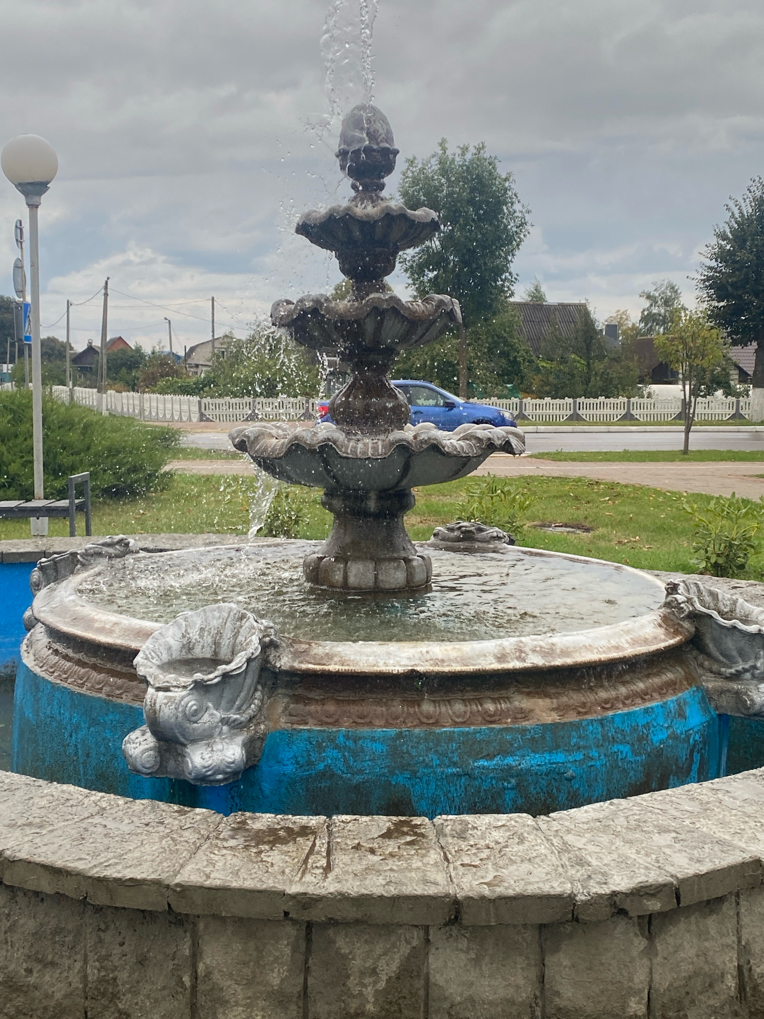 The image shows a multi-tiered fountain with water flowing from the top. The fountain is made of stone and has decorative elements, including what appear to be fish heads spouting water around the base. The fountain is set in a paved area with grass and bushes surrounding it. In the background, there is a white picket fence, a road with a blue car, houses with tiled roofs, trees, and utility poles. The sky is overcast with clouds. A street lamp is visible to the left of the fountain. The scene is located in the city of Krupki, Belarus, and appears to be a public space, possibly a park or town square. There are no people visible in the image.