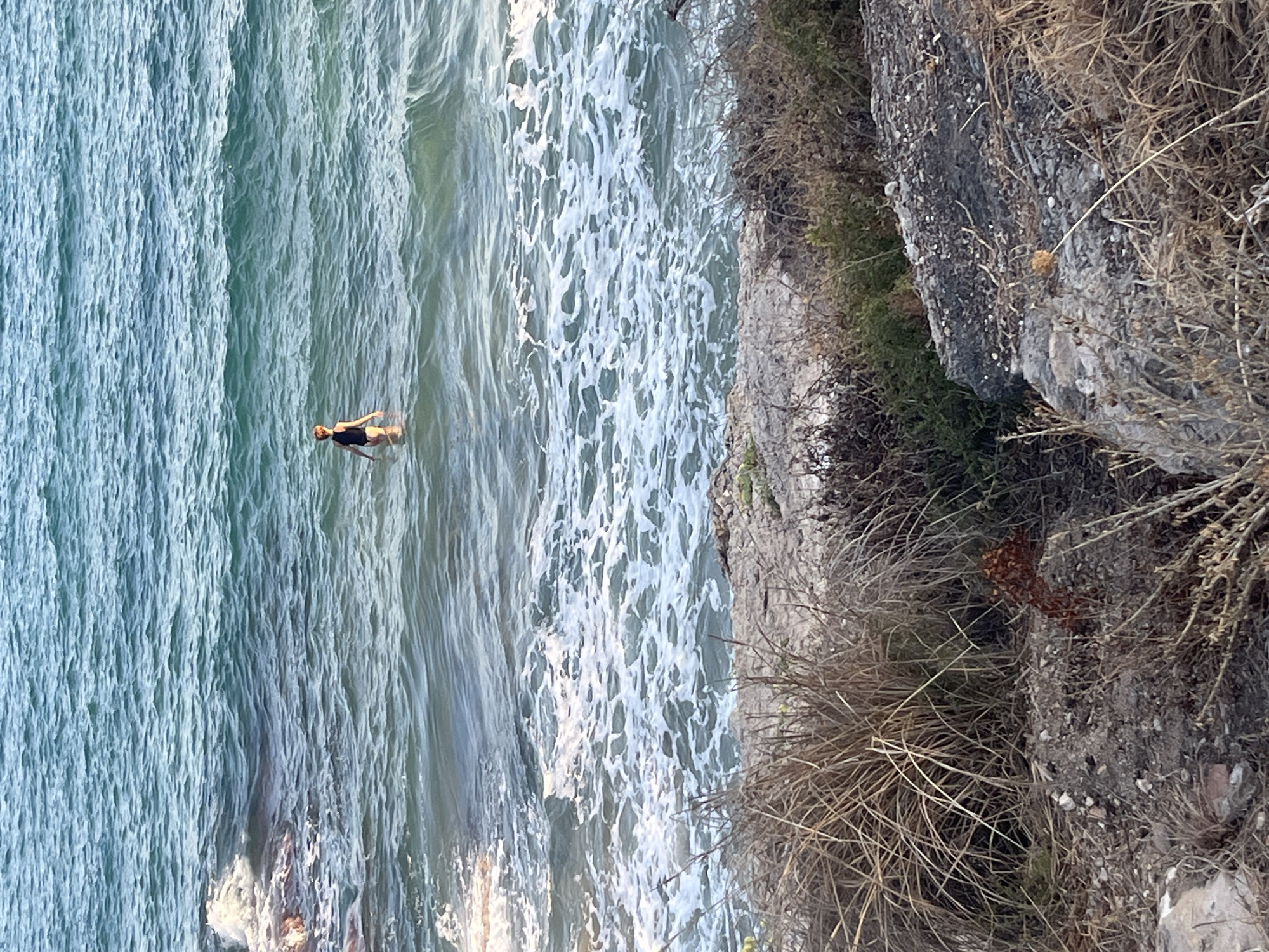 A lone person, appearing as a child, stands in the shallow waters of the sea at El Miracle beach in Barcelona, Spain. The individual is wearing a black swimsuit. The water is a deep blue-green, with visible waves and foam near the shore. The foreground is dominated by dry, brown vegetation and rocks, suggesting a somewhat unkempt, natural coastal area. The lighting indicates it is likely daytime, with the sun casting a natural glow on the scene.