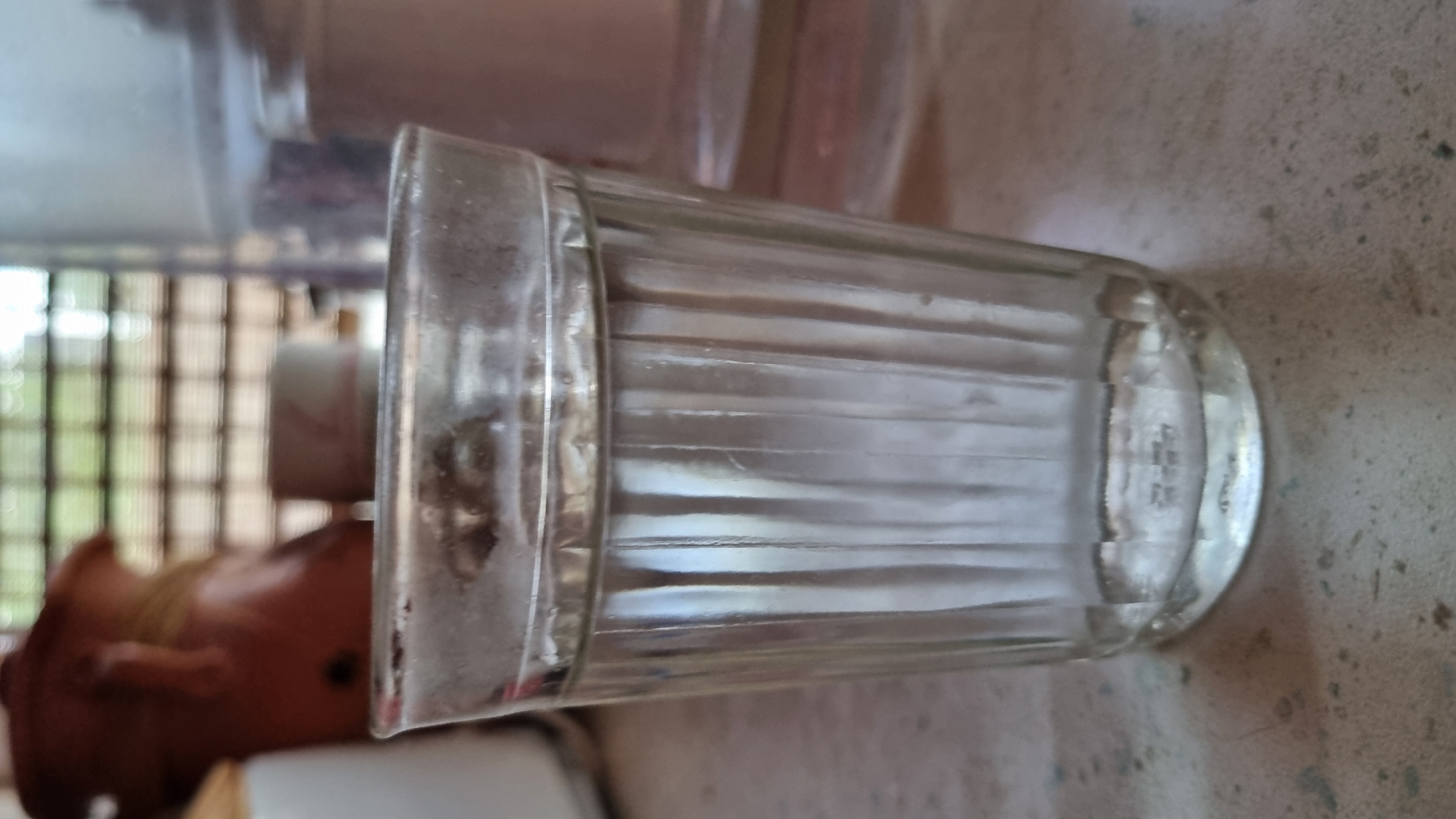 A close-up shot shows a clear glass with vertical fluting, filled with a clear liquid, sitting on a speckled surface. The glass is positioned in the foreground, with its rim slightly fogged. In the background, out of focus, a brown, amorphous object rests to the left, and a cylindrical, translucent container is visible to the right. Behind these, a window with horizontal blinds is apparent, allowing diffused light to enter. The overall scene appears to be indoors, potentially in a kitchen or dining area. The location is identified as Солнечная Поляна-3 Снт, Russia.