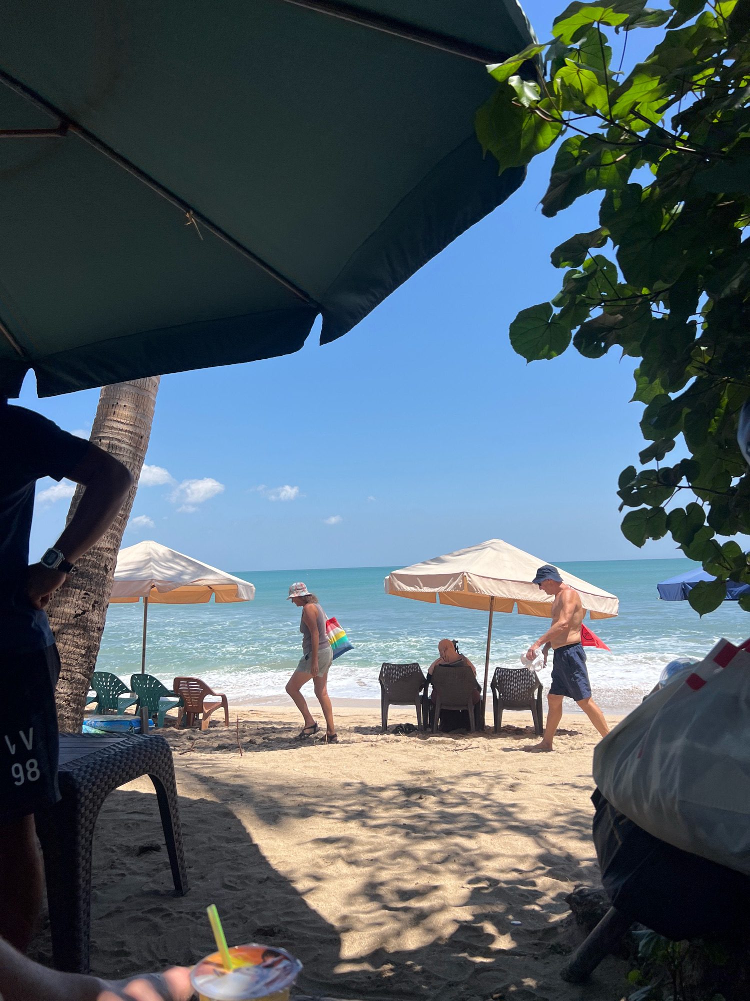 In Kuta, Indonesia, a beach scene unfolds under a bright, sunny sky. The foreground is partially obscured by a large, dark green umbrella and the trunk of a palm tree. A person's arm, wearing a dark t-shirt with white lettering and numbers "LV 98", is visible on the left, holding a beverage with a yellow straw. To the right, a portion of a person in the foreground is visible, wearing dark shorts and a tan bag with red and white accents.
The middle ground features a sandy beach with several beach chairs and umbrellas. A light-colored umbrella with a reddish border is set up, and two people are seated in chairs beneath it. Further out, a woman wearing a hat and a colorful bag walks towards the water, where waves are gently breaking. Another man walks along the shoreline, wearing dark shorts and holding a white cloth. A blue umbrella is visible in the distance. The ocean stretches out to the horizon, meeting a clear blue sky dotted with a few white clouds. Green foliage from a tree is visible in the upper right corner, casting shadows on the sand.